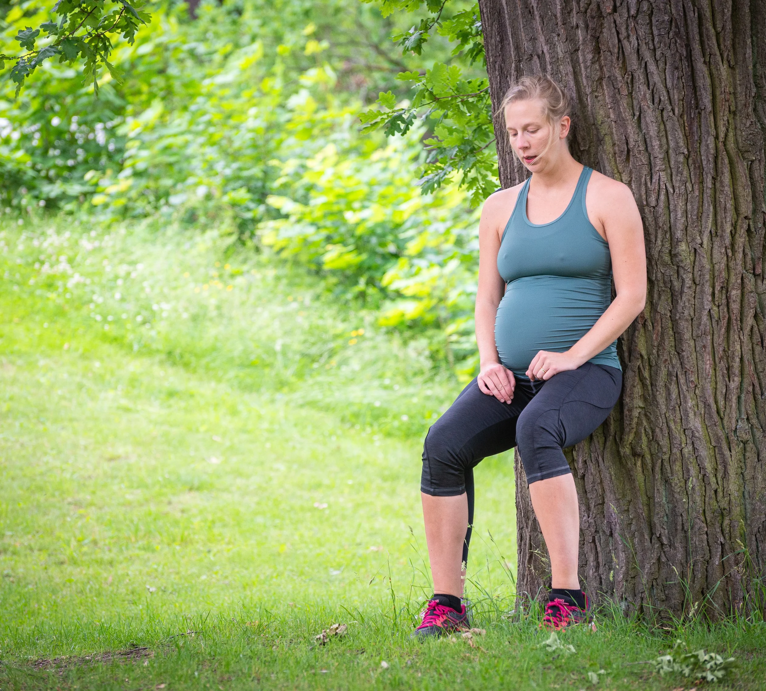 En gravid kvinna står lutad mot ett träd i en grön park, iklädd träningskläder och rosa sportskor.