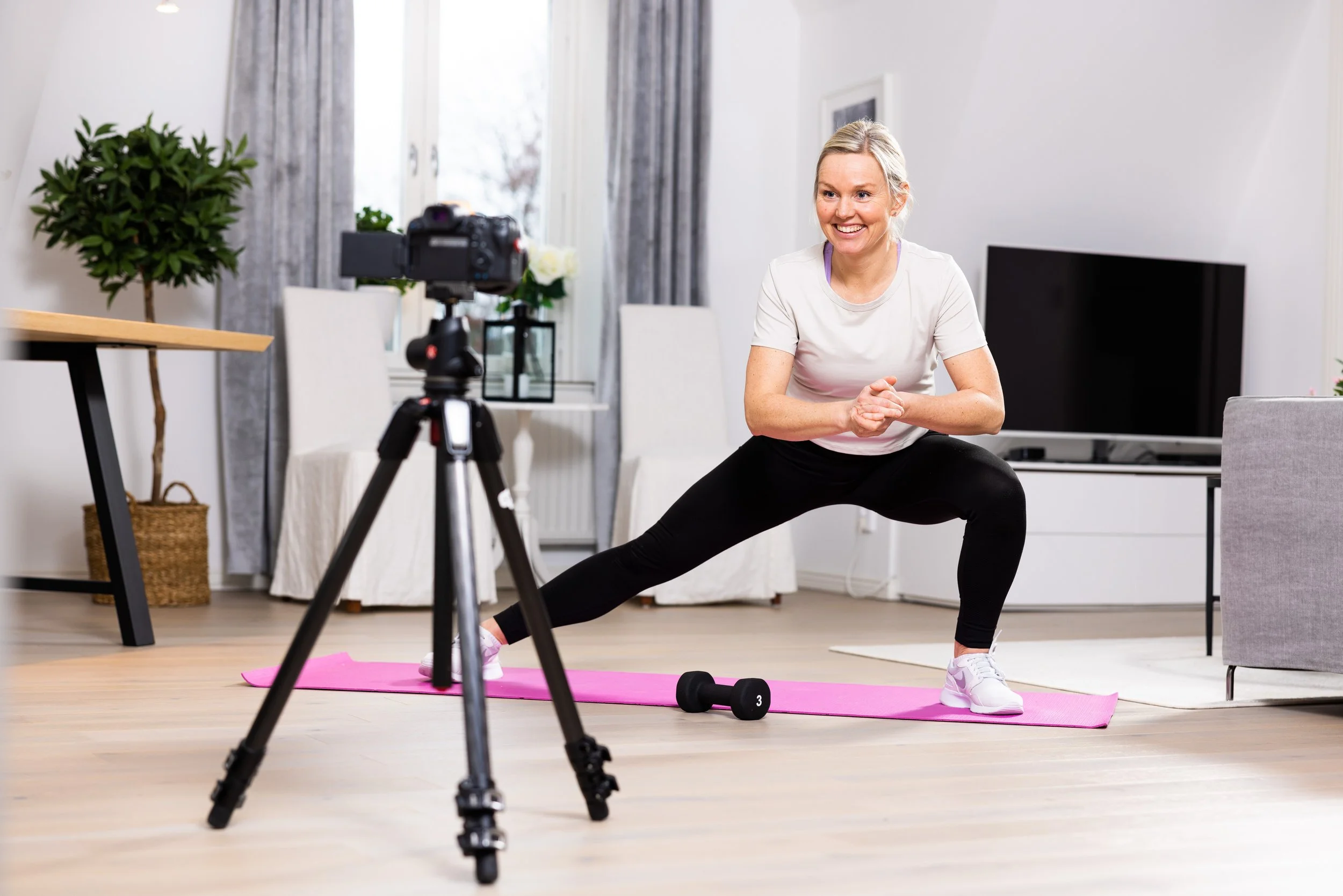En kvinna som tränar yoga i ett hem, utför en bredstående squat på en rosa träningsmatta, med en kamera på stativ riktad mot henne.