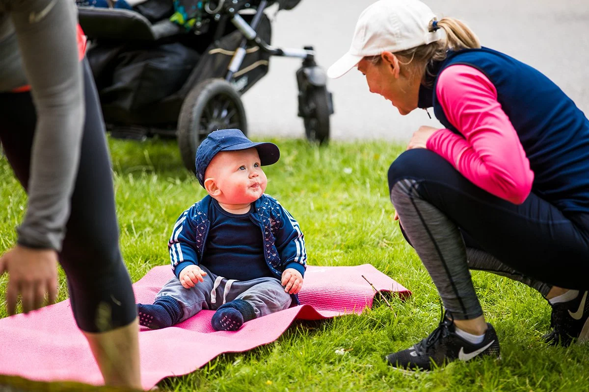 En baby sitter sittande på en rosa matta på gräset, prata med en liten baby som sitter framför hen, och en person med bara benen synliga till vänster. Baby bär keps och träningskläder, och kvinnor har sportkläder.