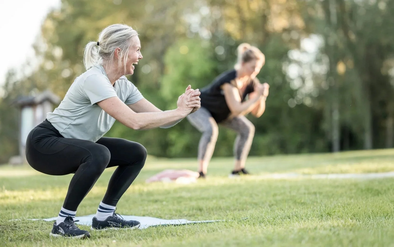 Två kvinnor gör squatövningar utomhus på gräs, med träd i bakgrunden, avkopplade och fokuserade.