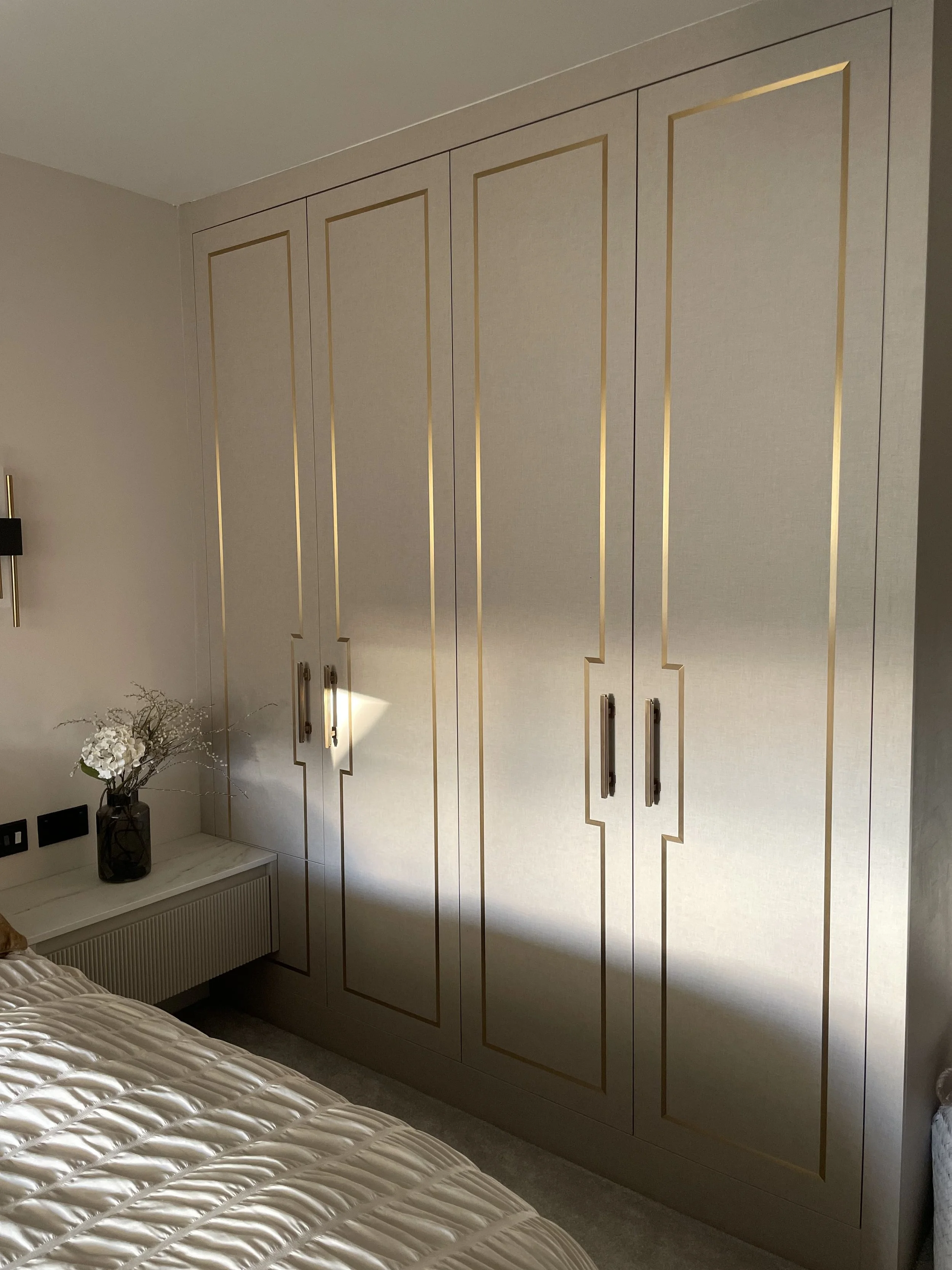 A modern bedroom featuring a large beige wardrobe with gold trim and handles, a bedside table with a black vase holding white flowers, part of a bed with a cream quilt, and a wall-mounted black and gold light fixture.