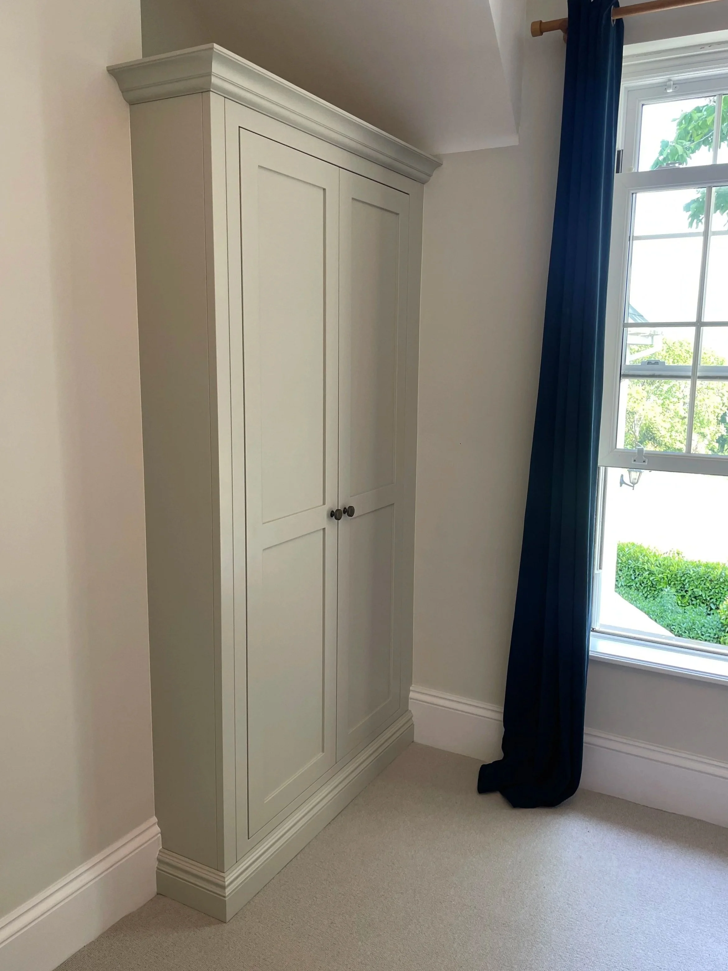 A white wardrobe with two doors and decorative molding in a corner of a room near a window with blue curtains. The room has beige carpet and white walls.