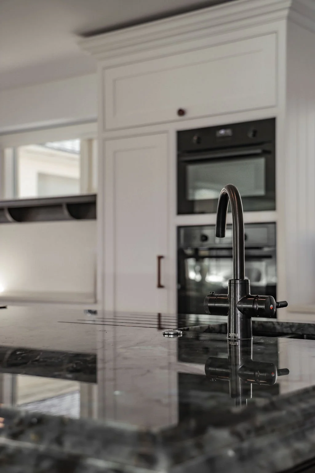 Close-up of a kitchen sink faucet with a black handle, granite countertop, with white cabinets and a built-in oven in the background.