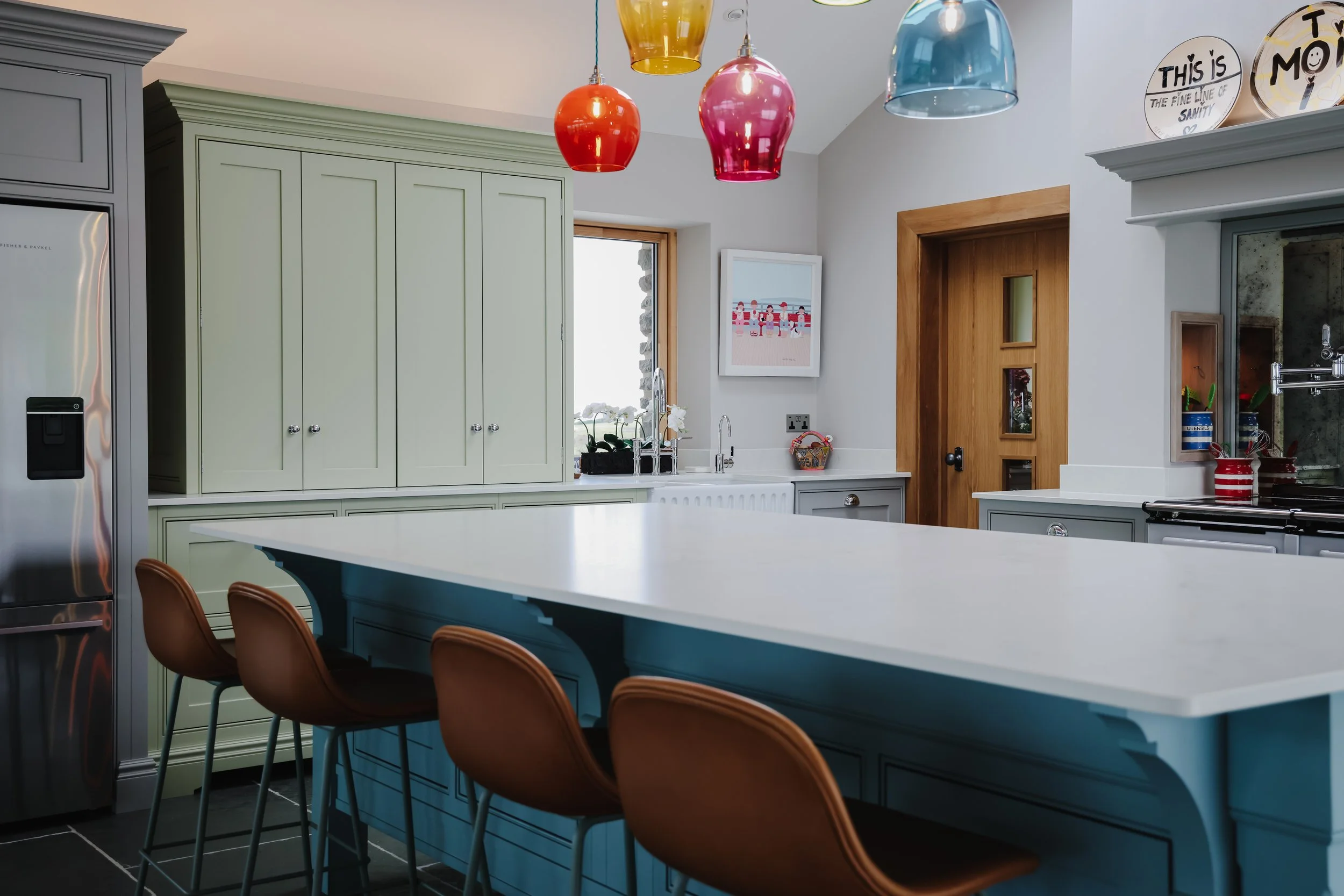 Modern kitchen with pastel green and blue cabinetry, a large white island, orange bar stools, colorful pendant lights, a window with plants, a wooden door, and wall decor.