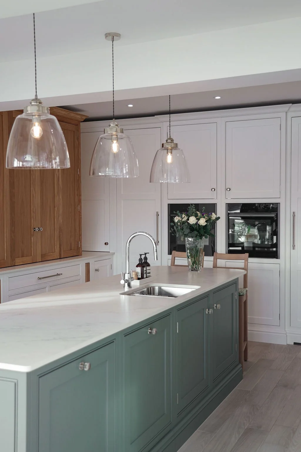 Modern kitchen with light green island, white cabinets, and wooden accents. Three pendant lights hang above the island, with a vase of white roses on the countertop.