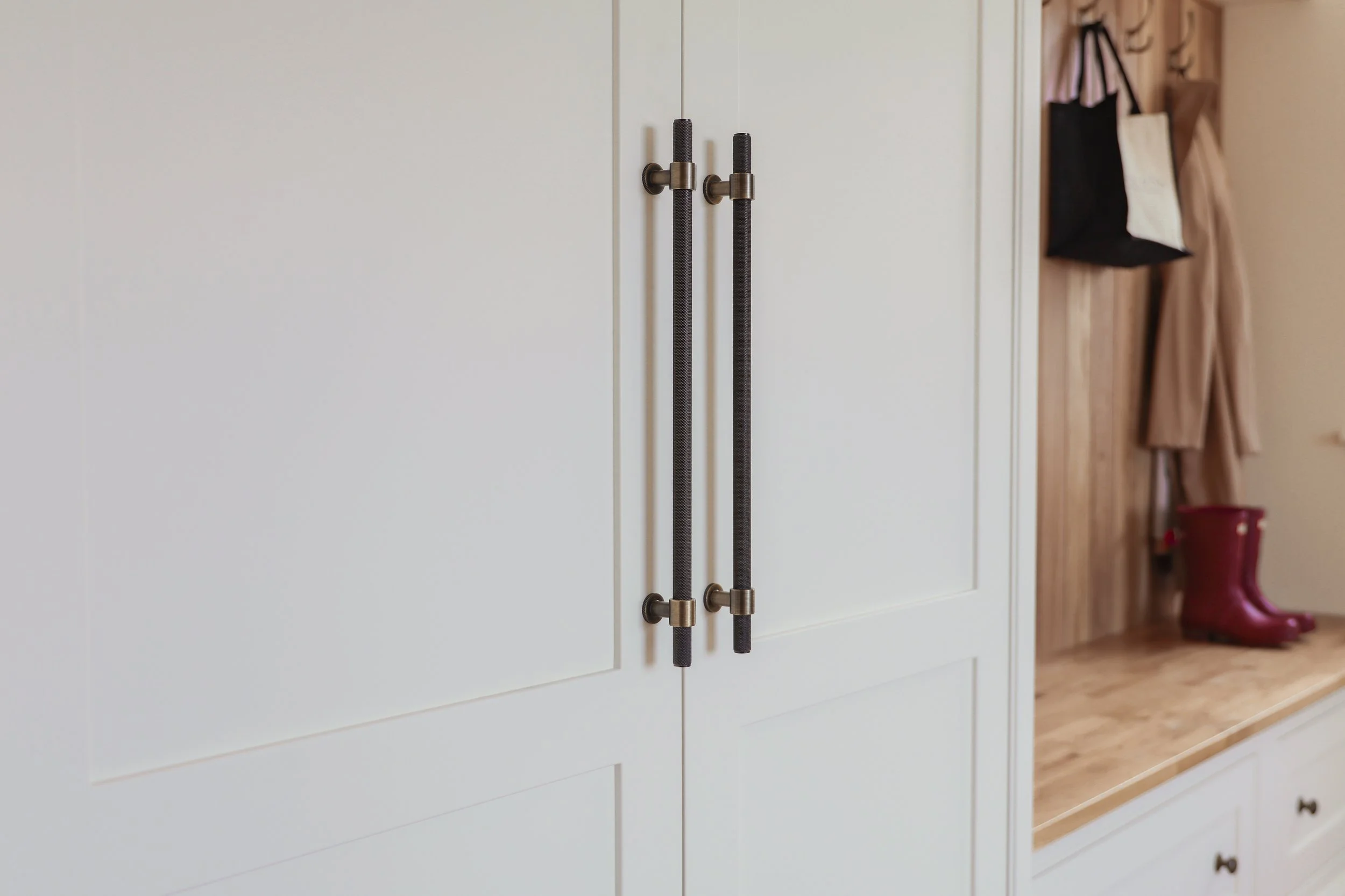 Close-up of white cabinet doors with black bar handles in a hallway, with a wooden shelf on the right holding a pair of burgundy rain boots, hanging bags, and coats.