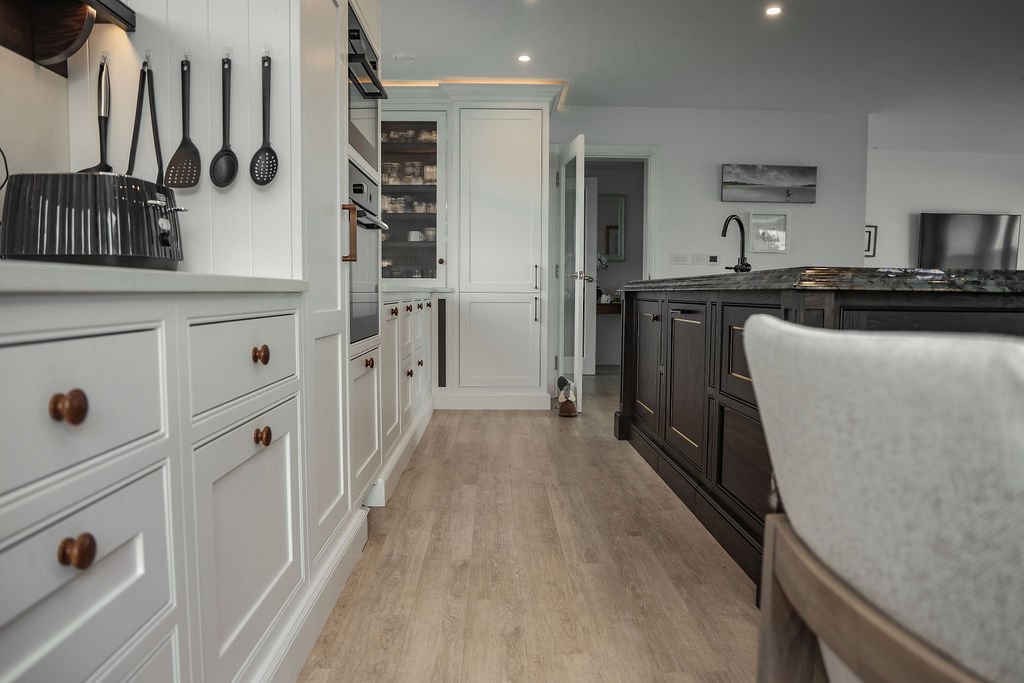 Modern kitchen with white cabinetry on the left, black utensils hanging on the wall, black toaster, and a dark island with a marble countertop on the right. Hardwood flooring and a doorway in the background.