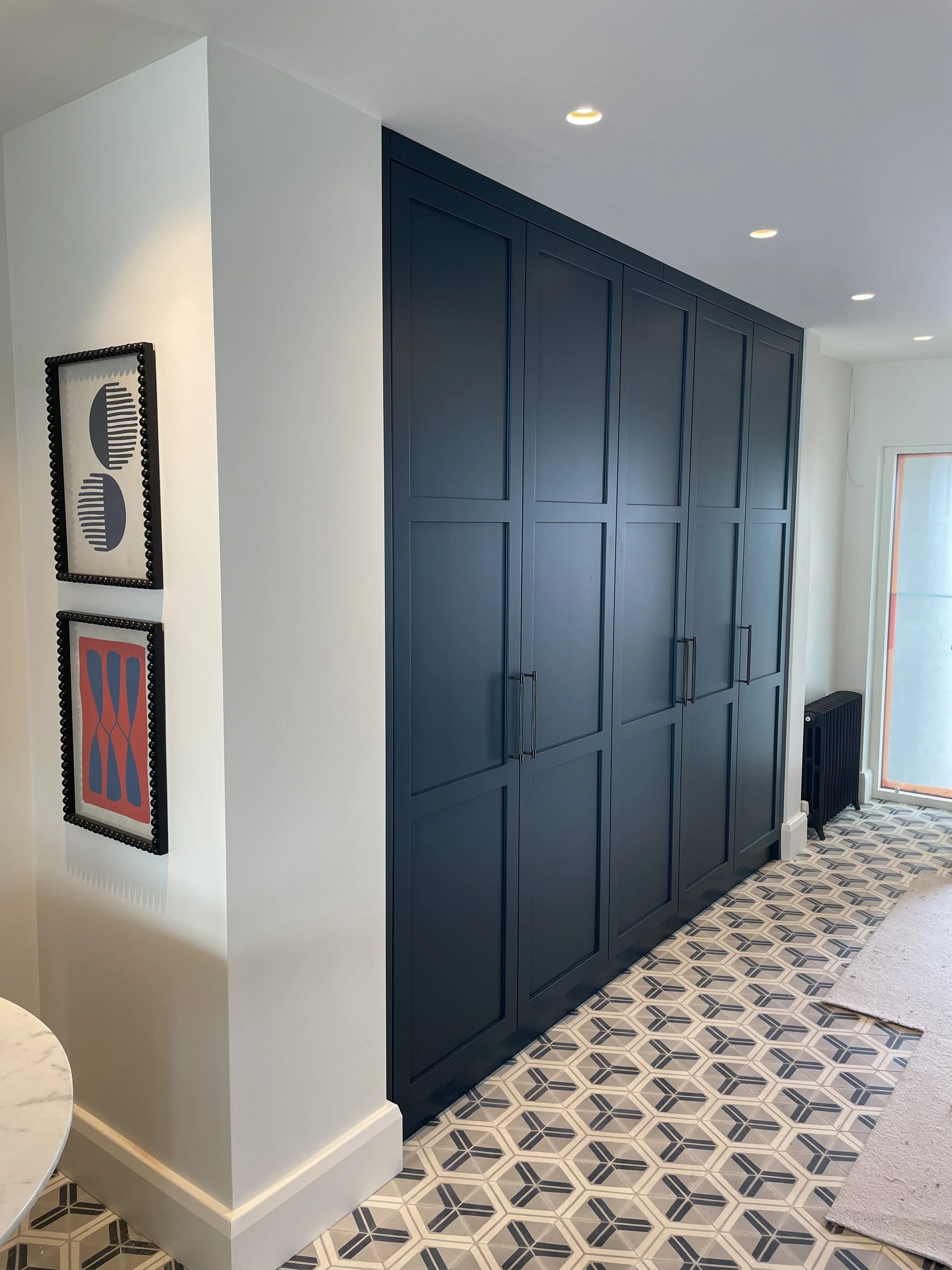 Dark blue built-in cabinets in a modern kitchen with patterned black and white tile flooring, white walls, recessed ceiling lights, a window, and a radiator.