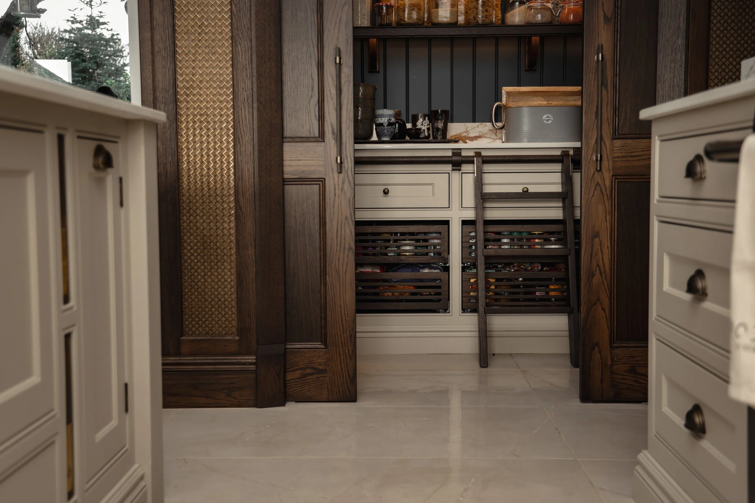 A view into a wine cellar with dark wood and white cabinetry, filled with wine bottles and glasses, with two wooden ladders leaning against the wine racks.