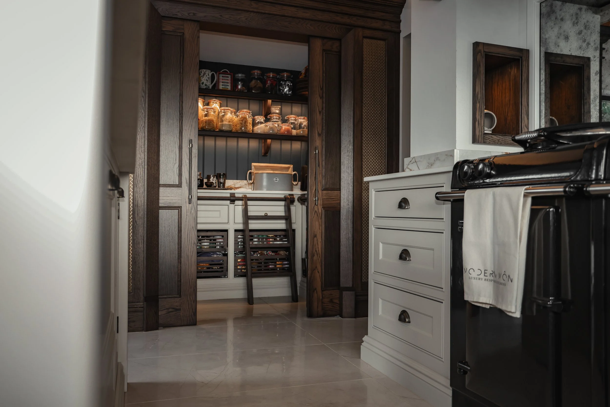 Pantry with open wooden doors showing shelves with jars of spices and dried goods, a white cabinet with drawers, and a black vintage stove with a dish towel hanging from its handle.