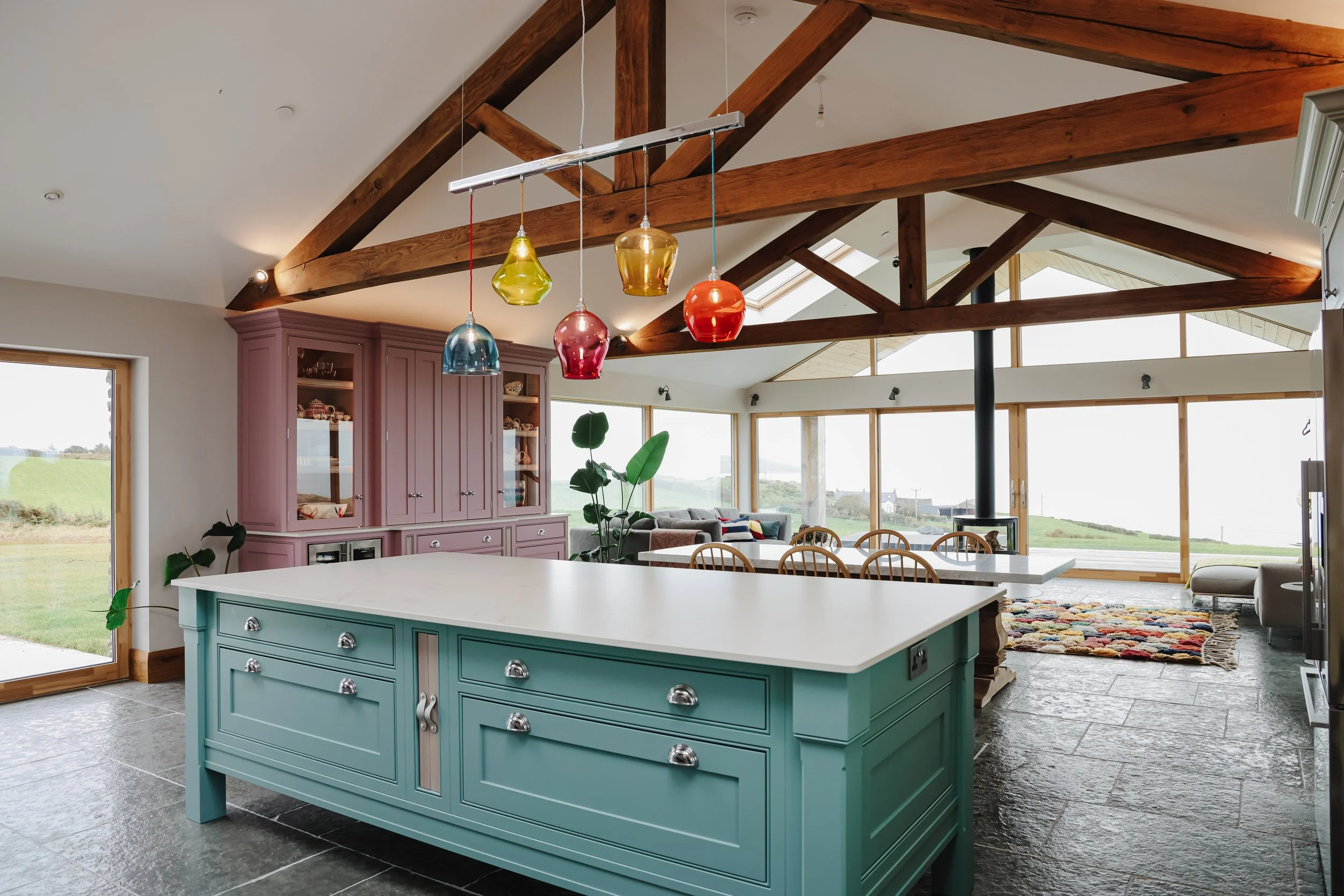 Interior of a spacious, modern home with a large kitchen island, colorful pendant lights, a cozy living area with a wood stove, and large windows showcasing a scenic outdoor view.
