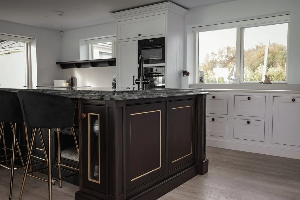 Modern kitchen with white cabinetry, large window, and a dark island with a marble countertop, bar stools, and built-in oven.