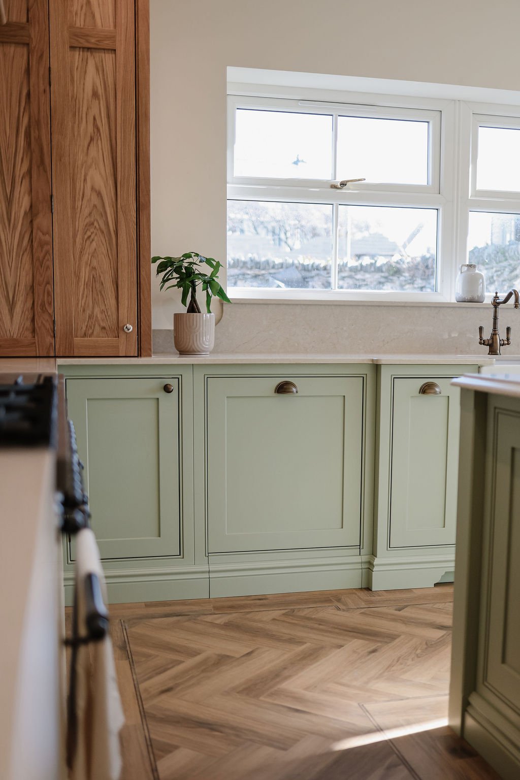 A cozy kitchen with green cabinetry, wooden upper cabinets, a window with a view, a potted plant on the countertop, and a herringbone wood floor.