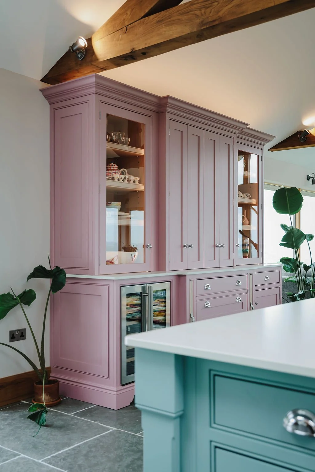 Pink kitchen cabinet with glass doors displaying dishes, set against a wall under a wooden beam ceiling, with green houseplants nearby and part of a teal-colored kitchen island visible in the foreground.