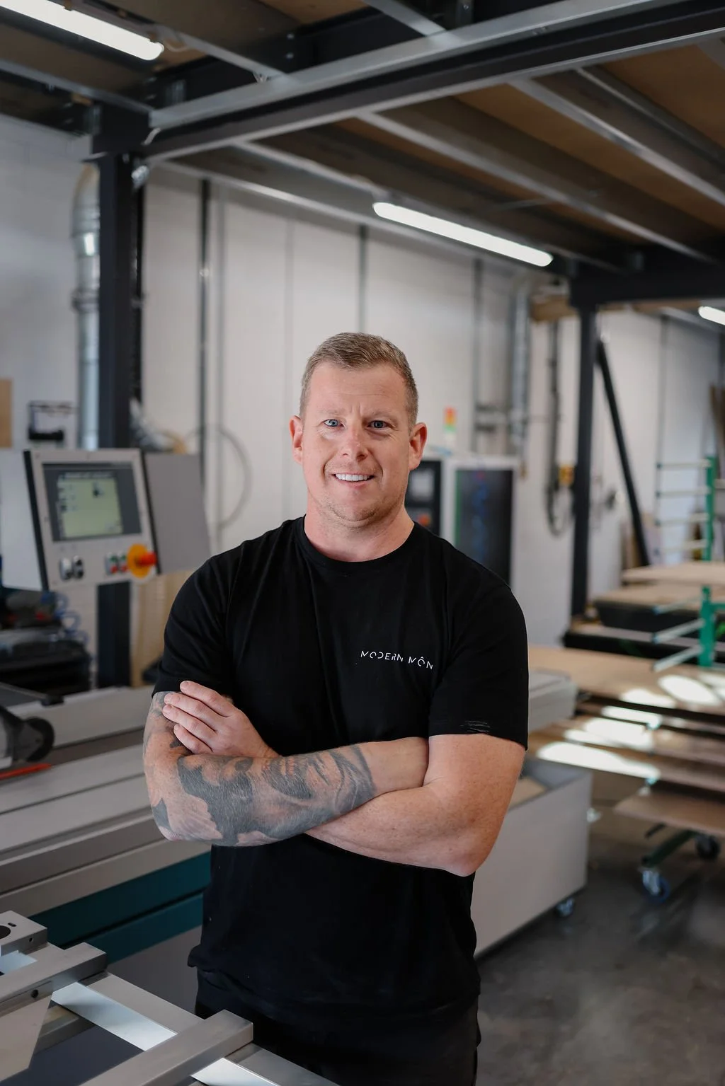 Man standing with arms crossed in woodworking workshop, wearing a black t-shirt, smiling, with woodworking machinery and materials in the background.