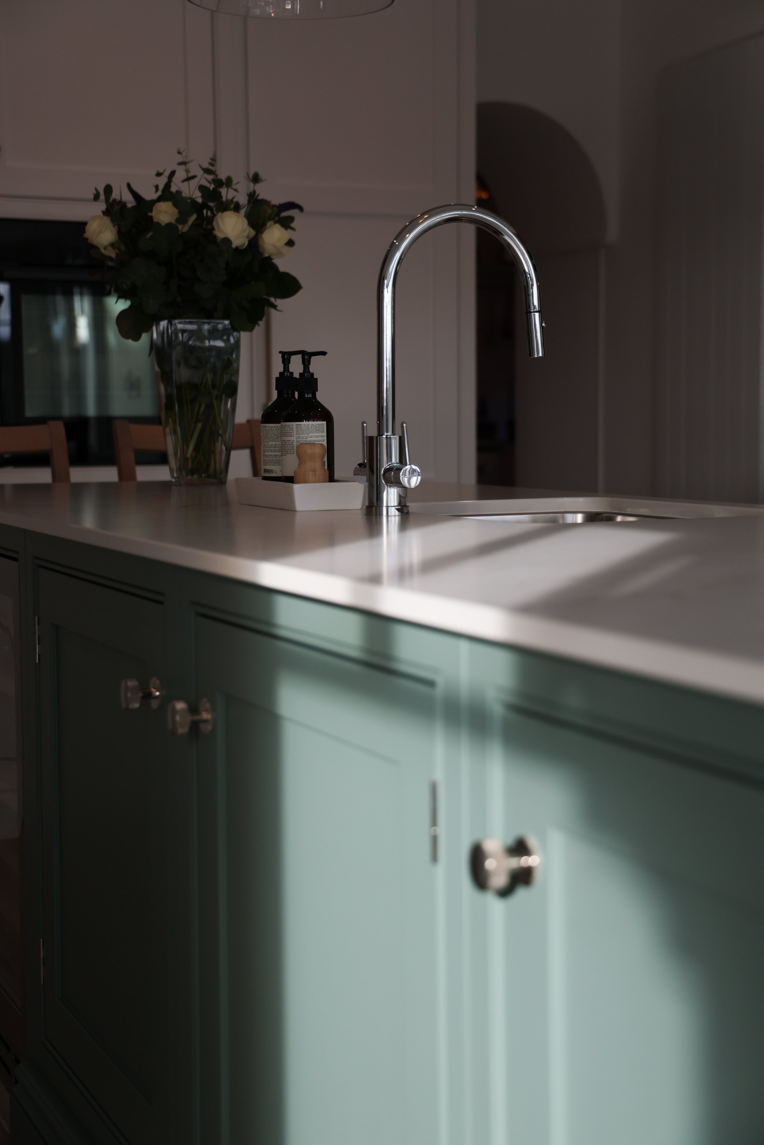 Kitchen island with a white countertop, a green lower cabinet, a faucet, a soap dispenser, and a vase of white roses in the background.
