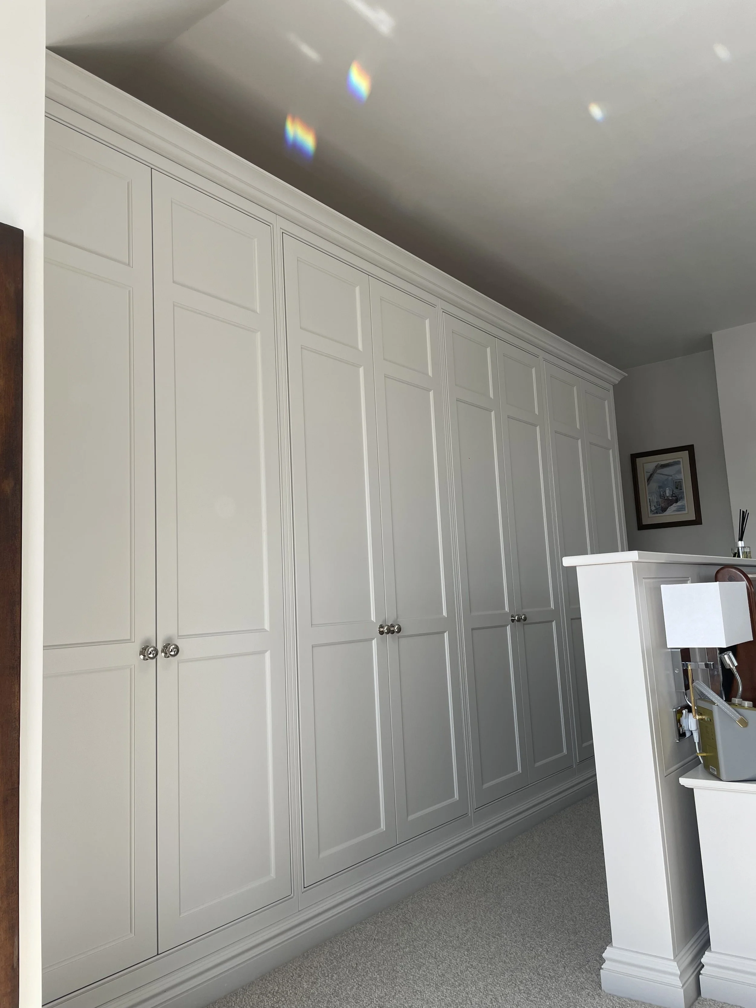 White built-in wardrobe with paneled doors and round knobs in a room with beige carpet and a wall-mounted picture frame.