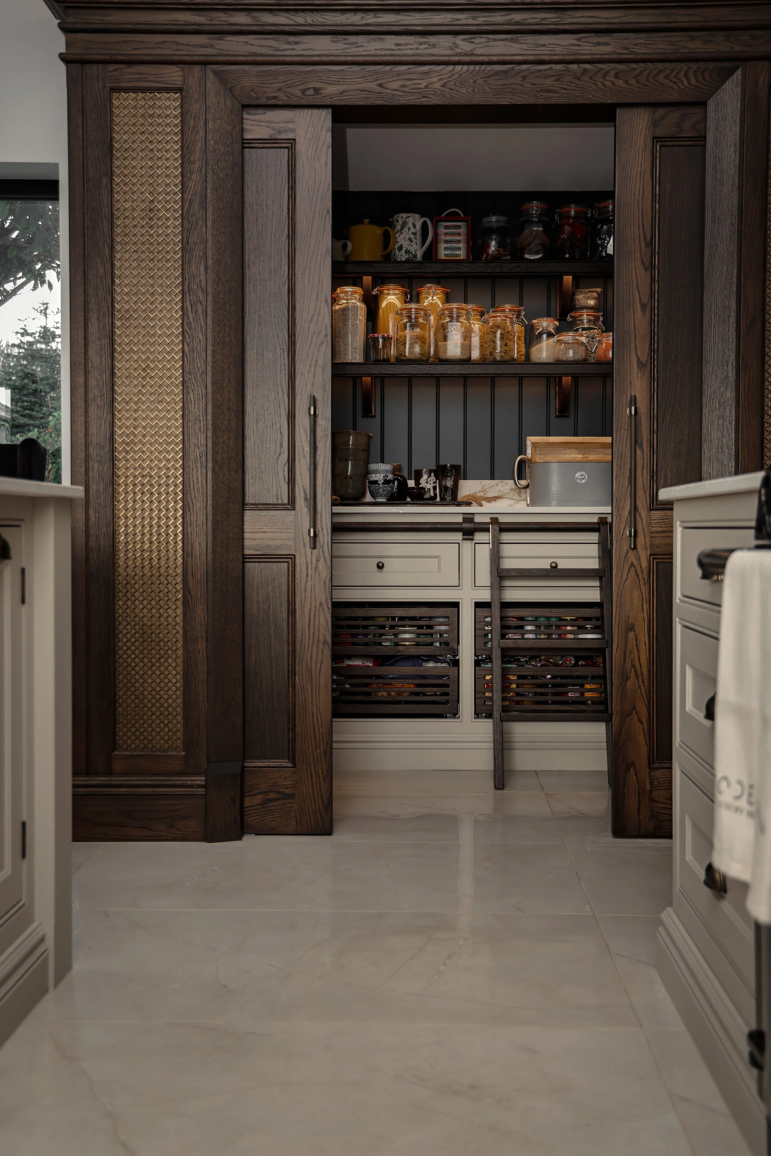 View of a kitchen pantry with open wooden sliding doors, showing shelves with jars and containers, a countertop, and kitchen utensils.