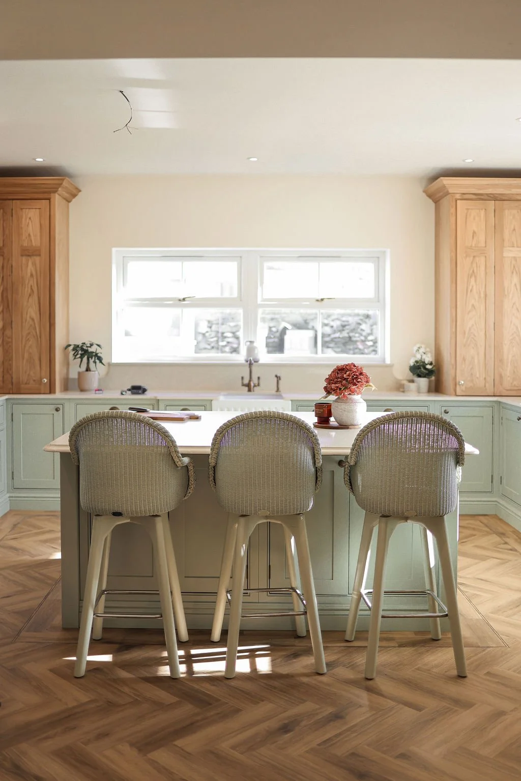 Modern kitchen with a central island, three upholstered bar stools, mint green cabinetry, wooden floors, and a window above the sink with potted plants and decorative items on the countertop.