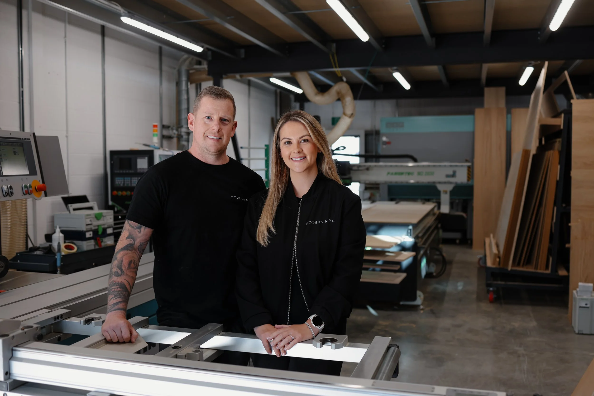 A man and woman standing in a woodworking workshop, smiling at the camera, surrounded by woodworking equipment and materials.