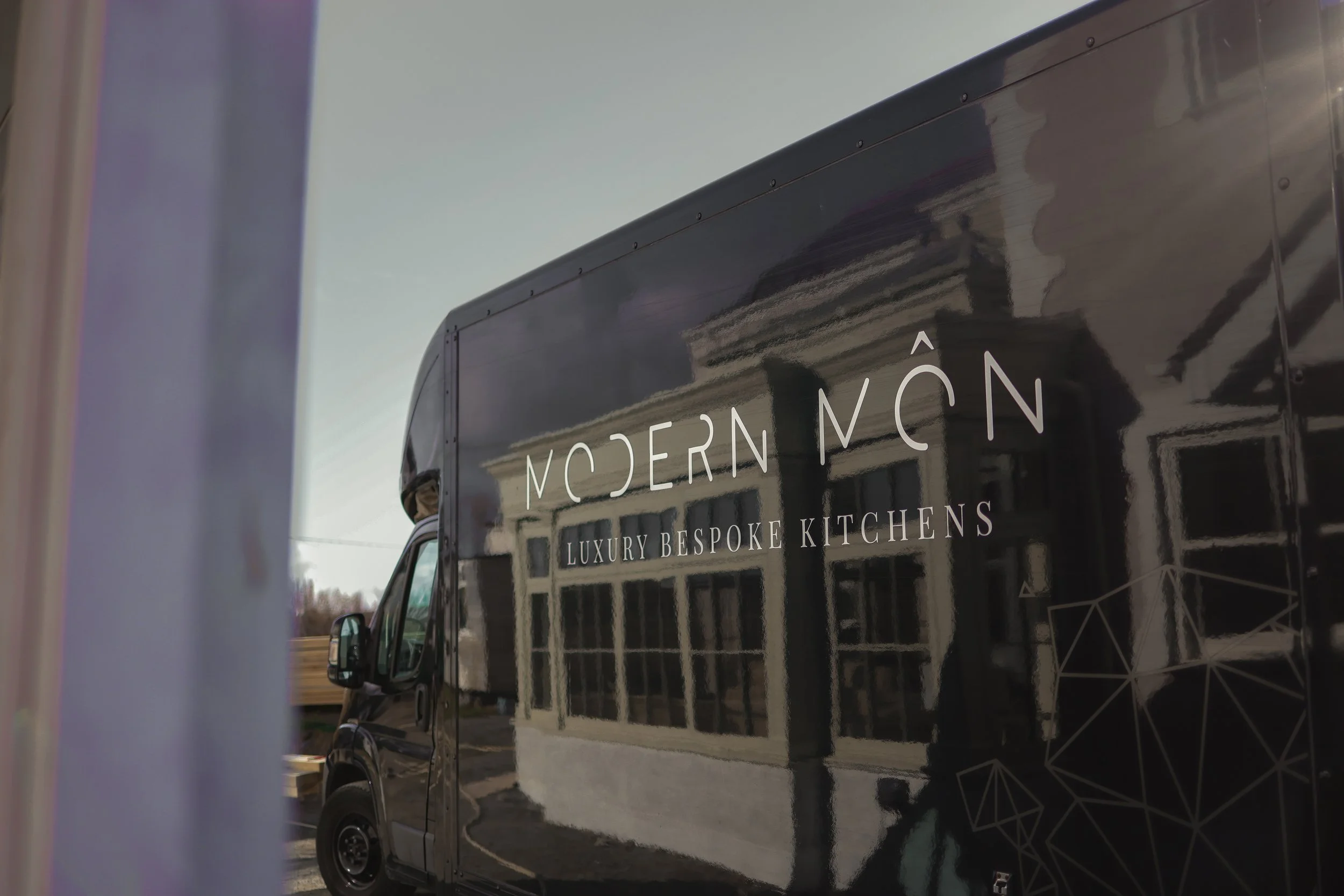 Black delivery truck with the logo 'Modern Mōn Luxury Bespoke Kitchens' reflected on its side, parked outdoors near a building with large windows.