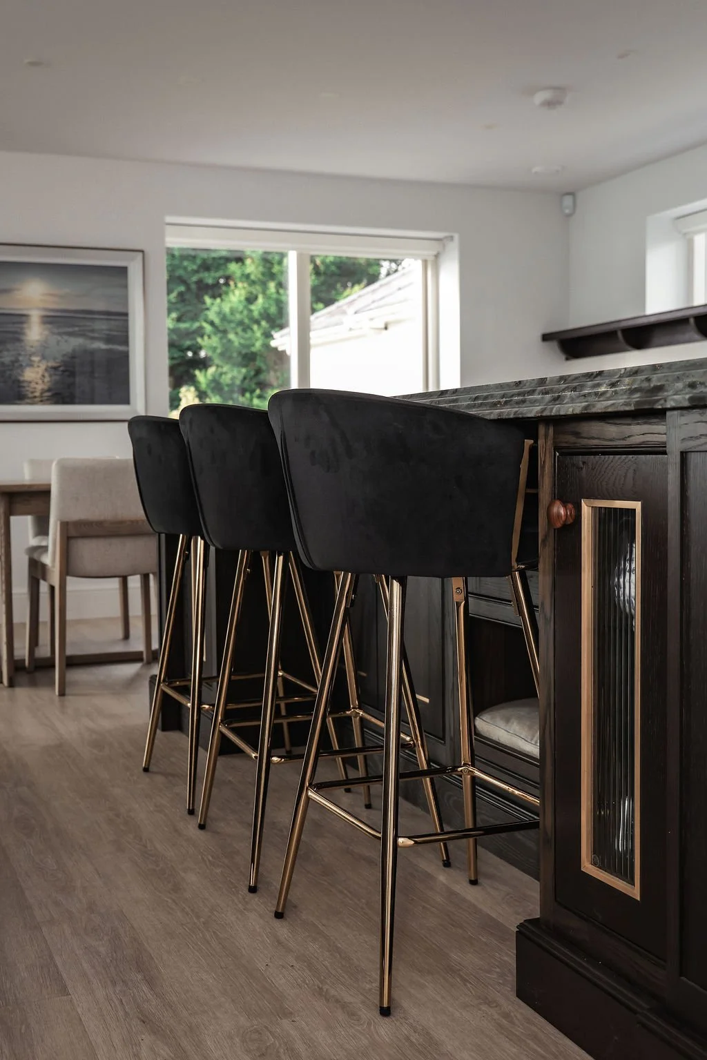 Kitchen with black barstools with gold legs at a dark wooden counter, and a beige dining chair in the background.