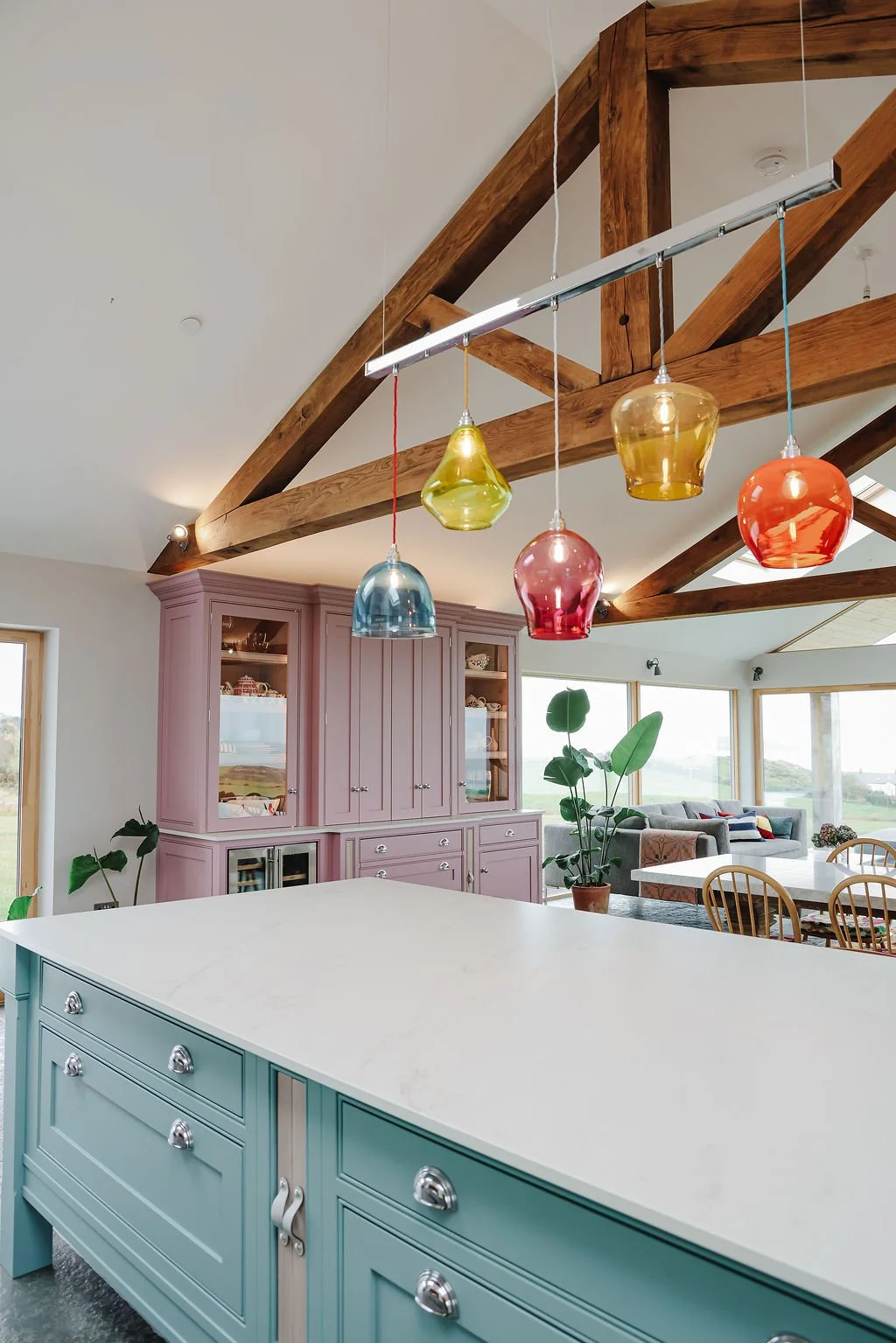 Inside a modern home with a kitchen island, pink cabinetry, colorful pendant lights, and large windows with a view of the outdoors.