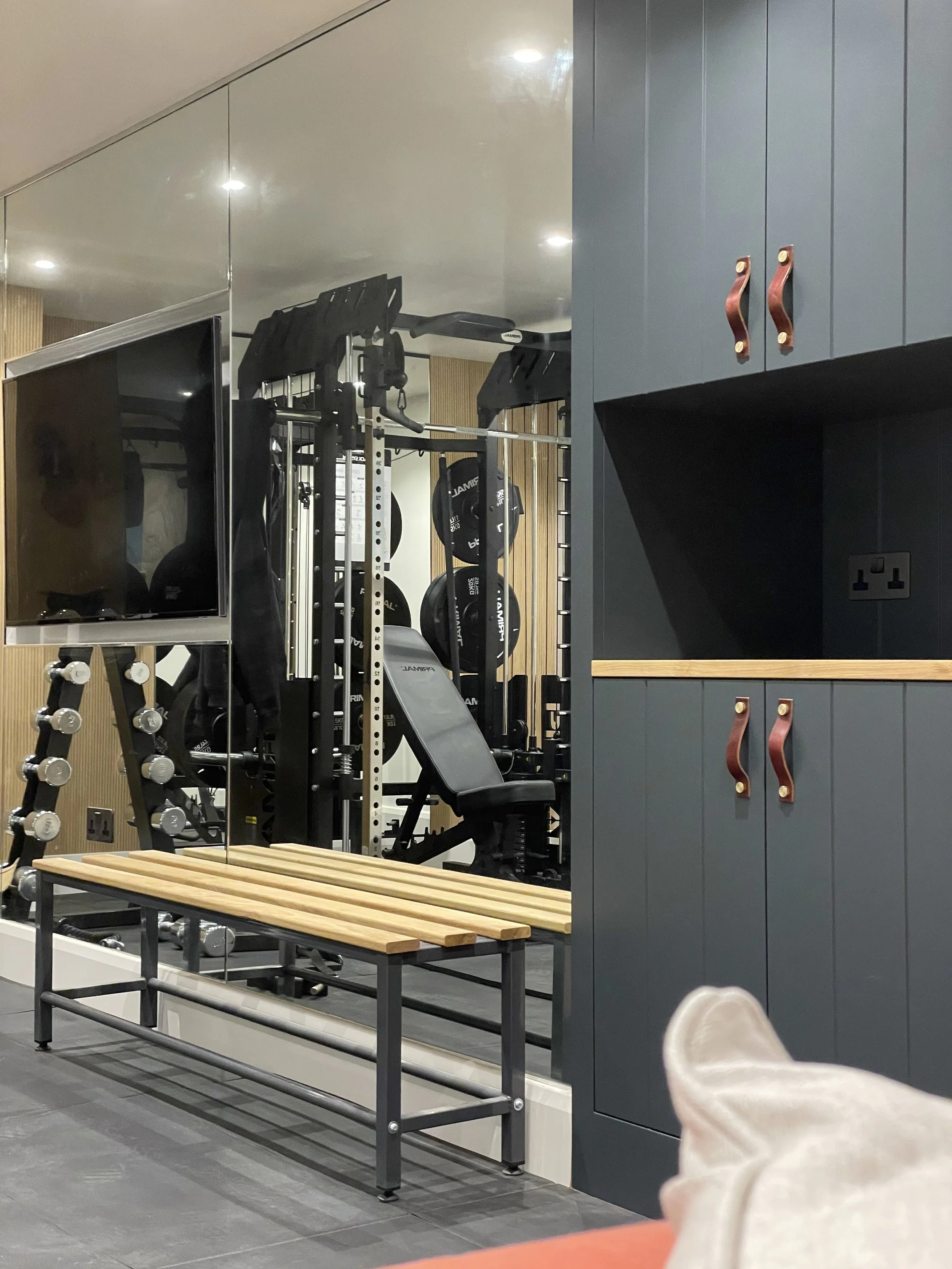 View of a gym with weightlifting equipment, including a bench press and dumbbells, seen through a mirror and separated by a glass wall, with a blue storage cabinet on the right side.