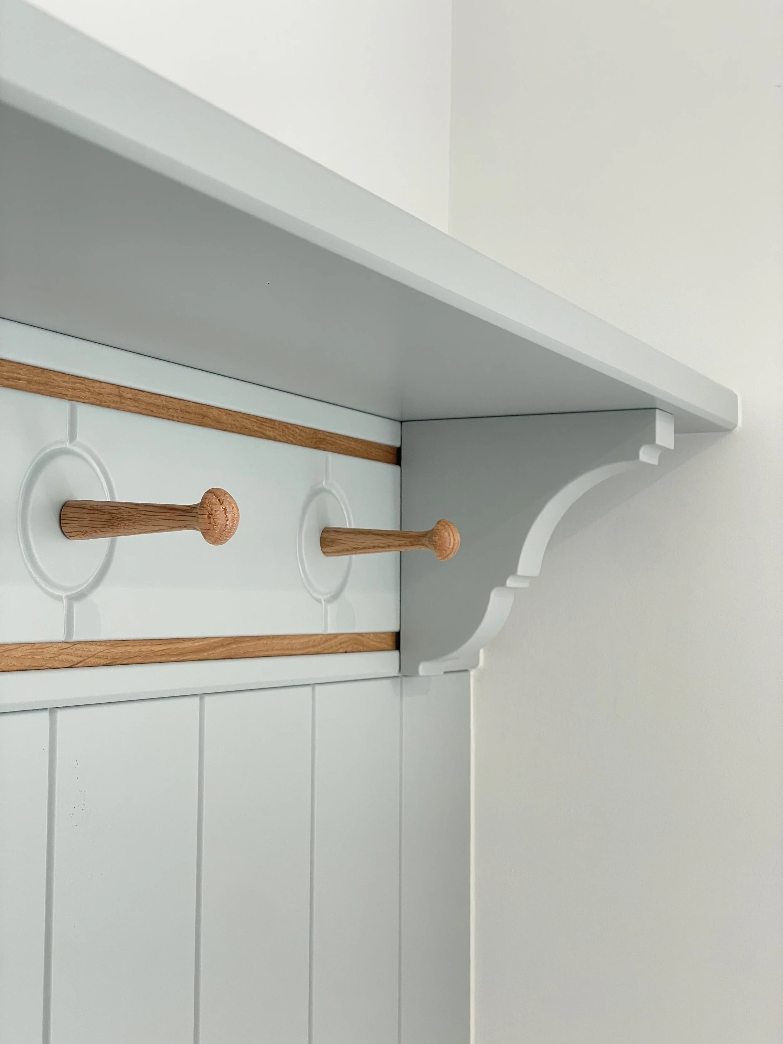 Close-up of a light-colored wooden coat rack with two pegs mounted on a white wall with vertical paneling, featuring decorative white trim.