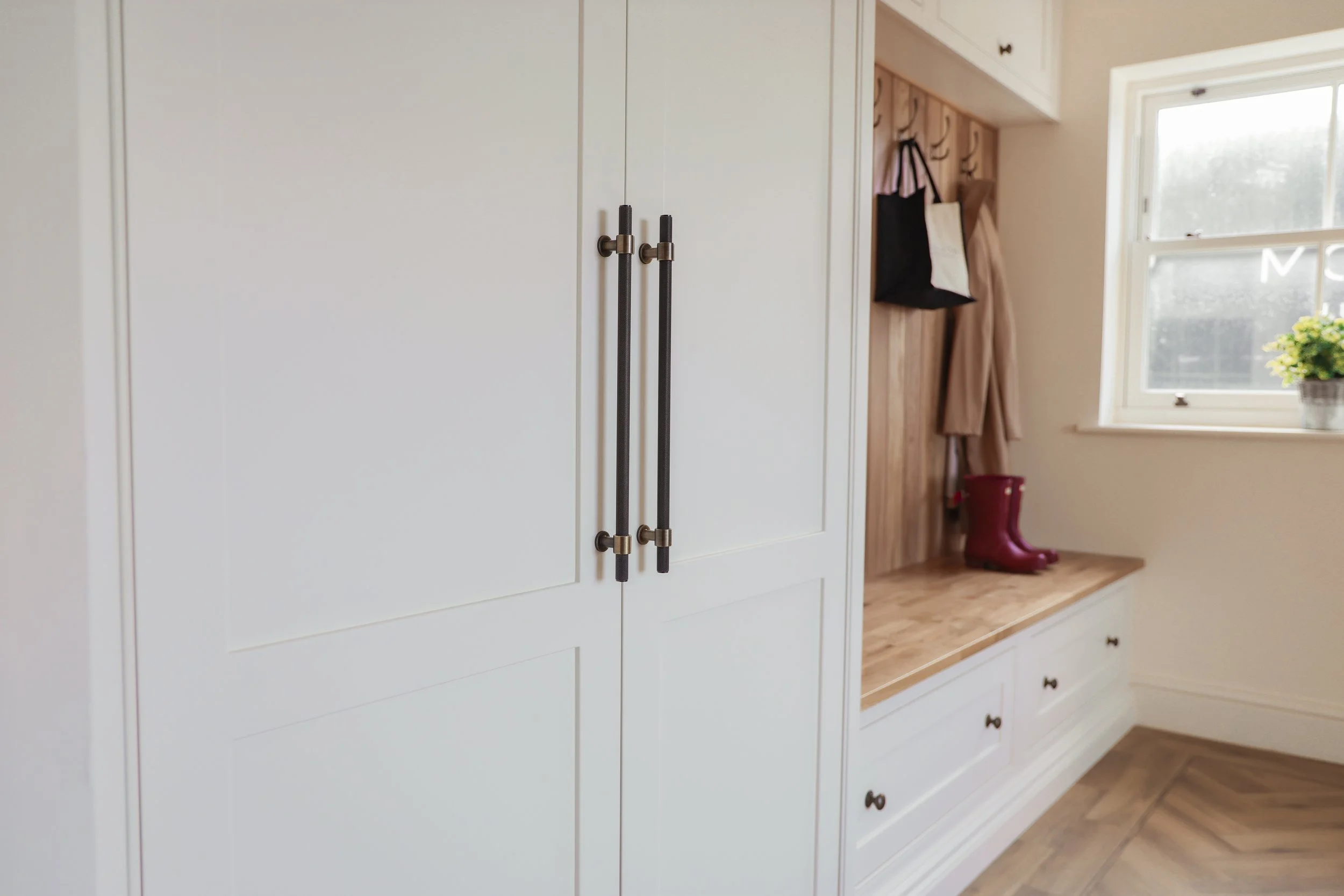 A cozy entryway with white built-in cabinets, dark handles, a wooden bench with drawers, and hooks holding bags, coats, and a pair of red boots. A window with a potted plant on the sill provides natural light.