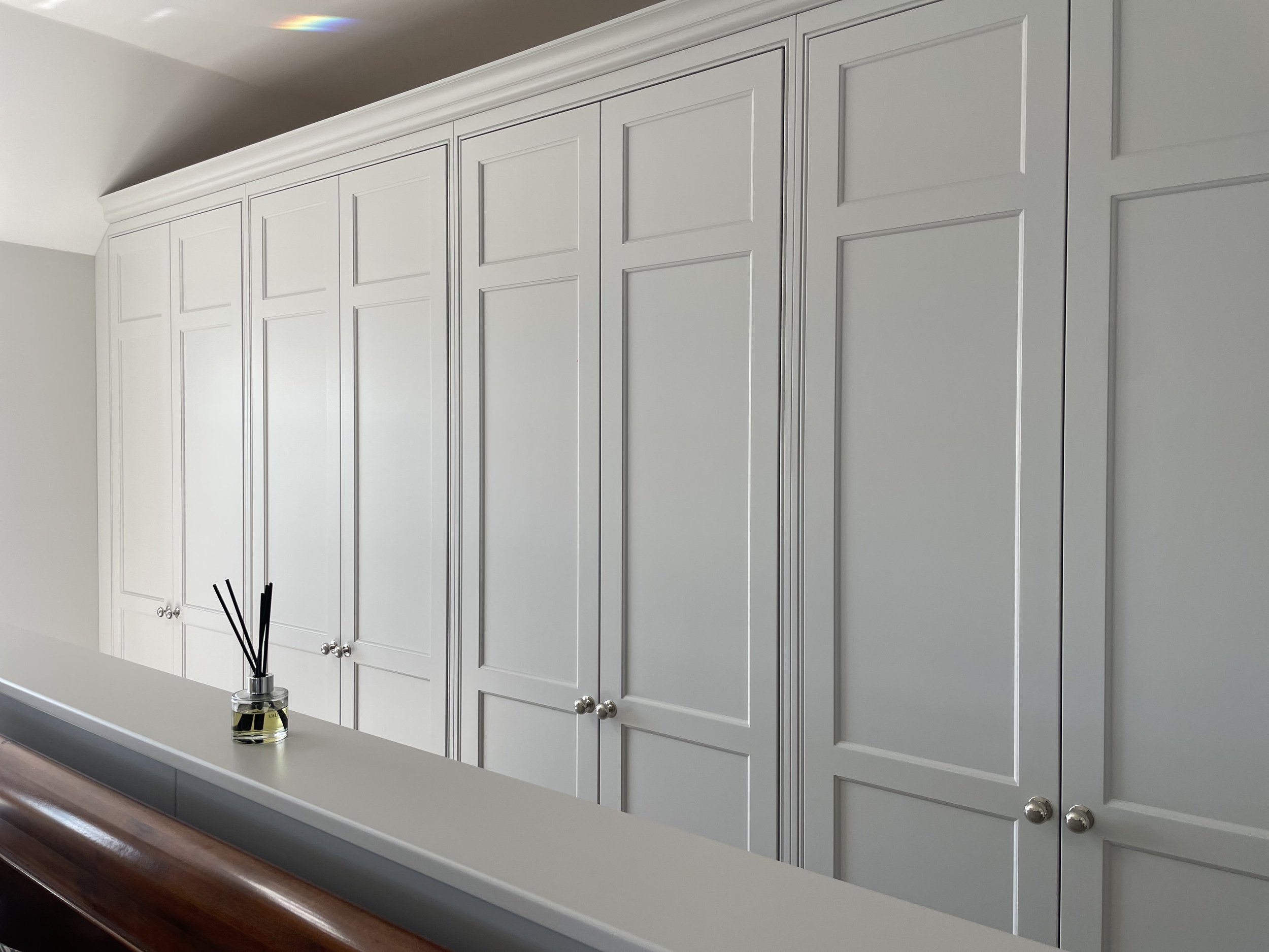 A row of tall white closet cabinets with panel doors and round knobs, next to a wall with a sloped ceiling, and a countertop with a reed diffuser in the foreground.