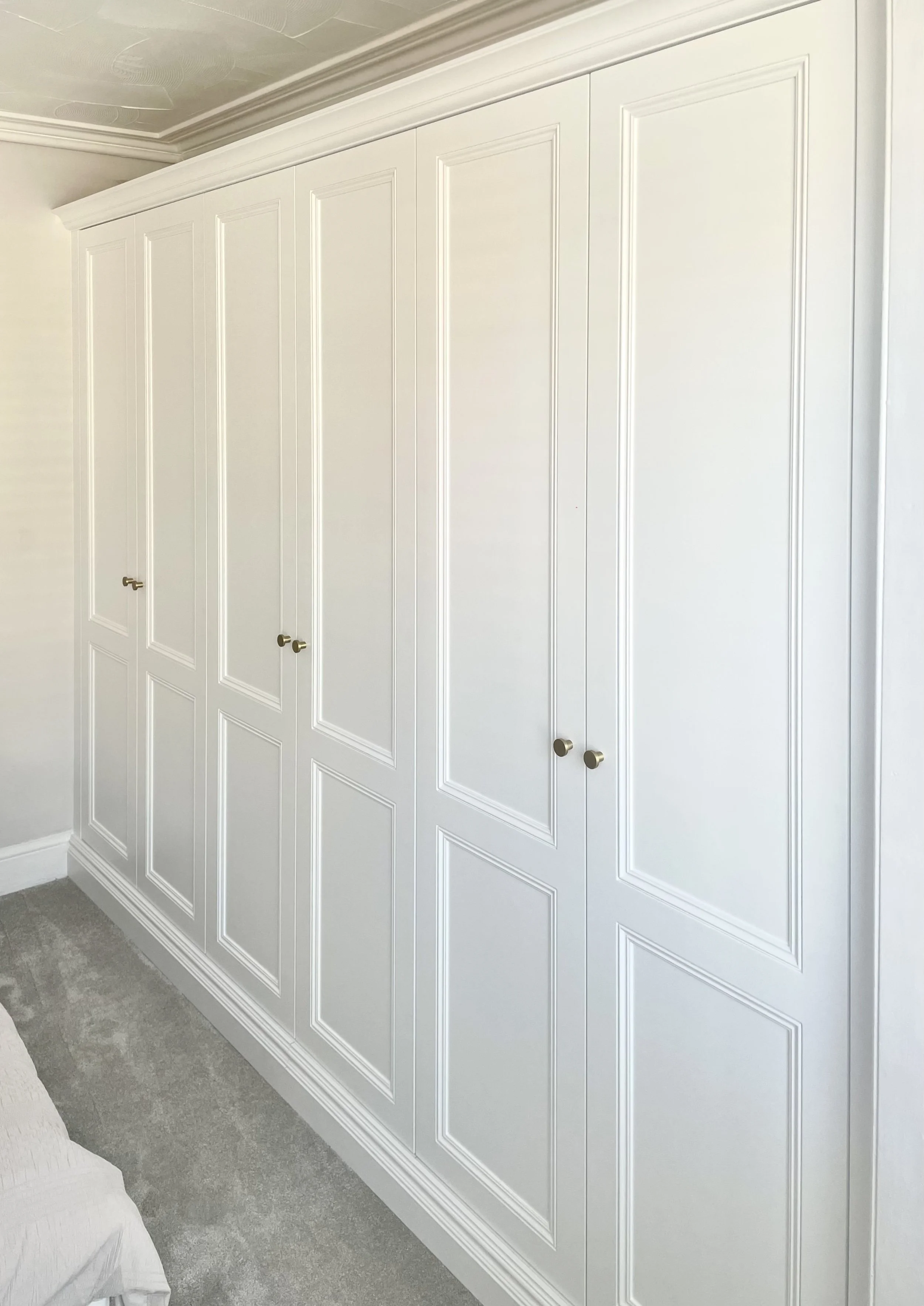 A large white built-in wardrobe with paneled doors and small brass knobs in a room with white walls and a light gray carpet.