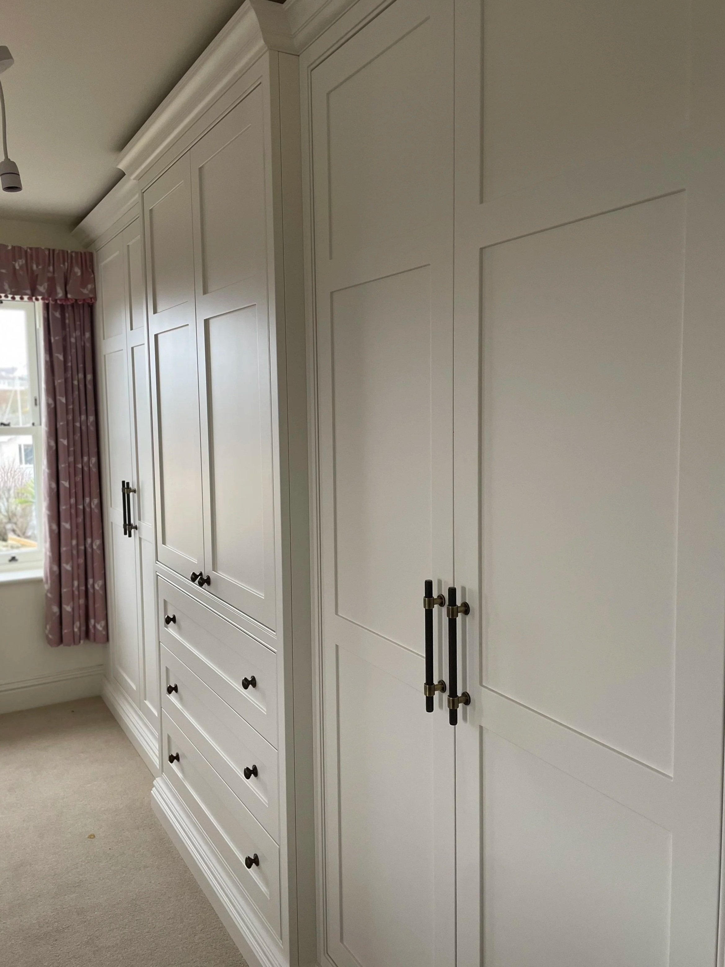 White built-in wardrobe with black handles in a room with beige carpet and a window with pink floral curtains.