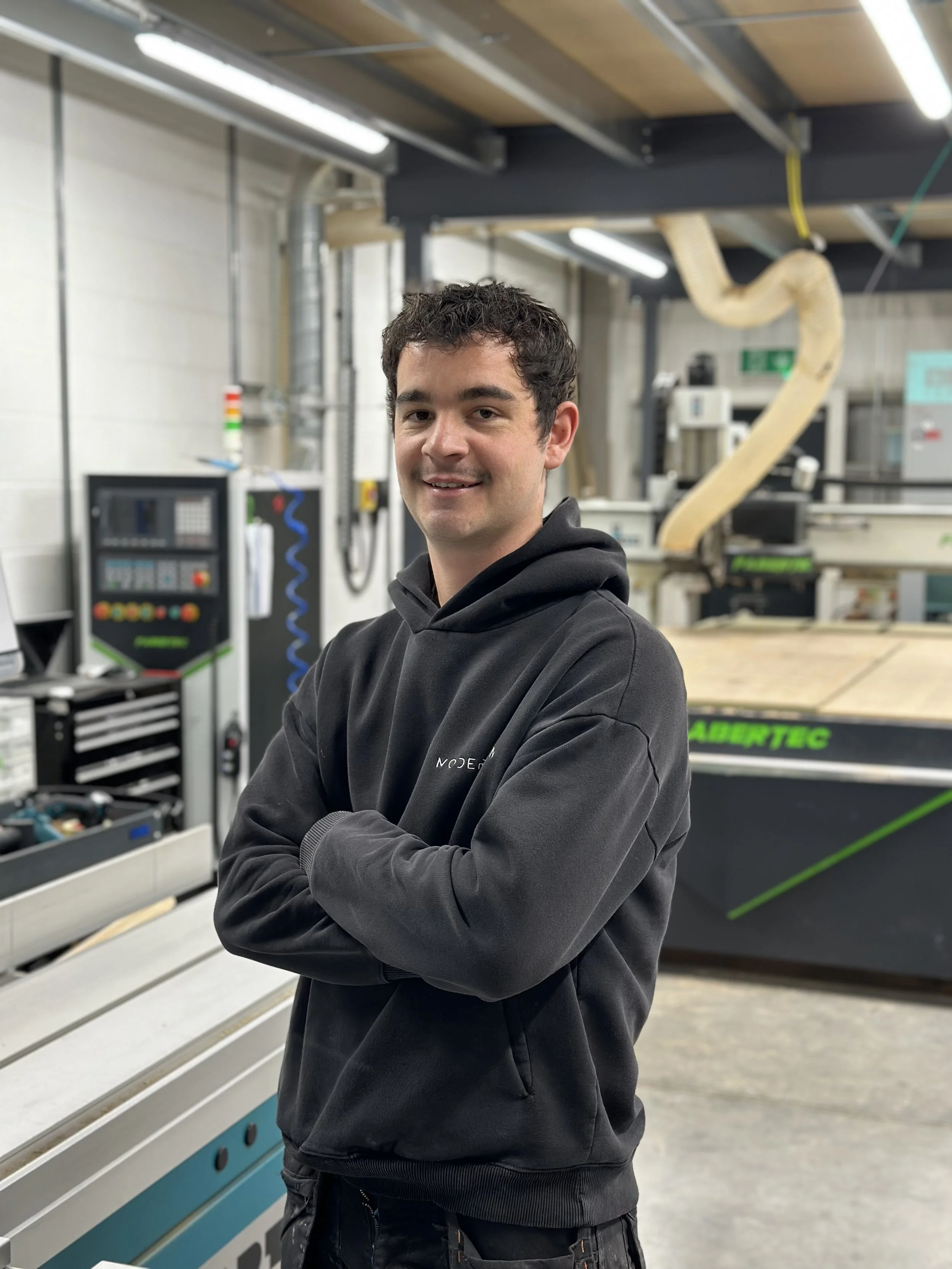 A young man with dark curly hair wearing a black hoodie, smiling with arms crossed, standing in a workshop or industrial setting with machinery and equipment.