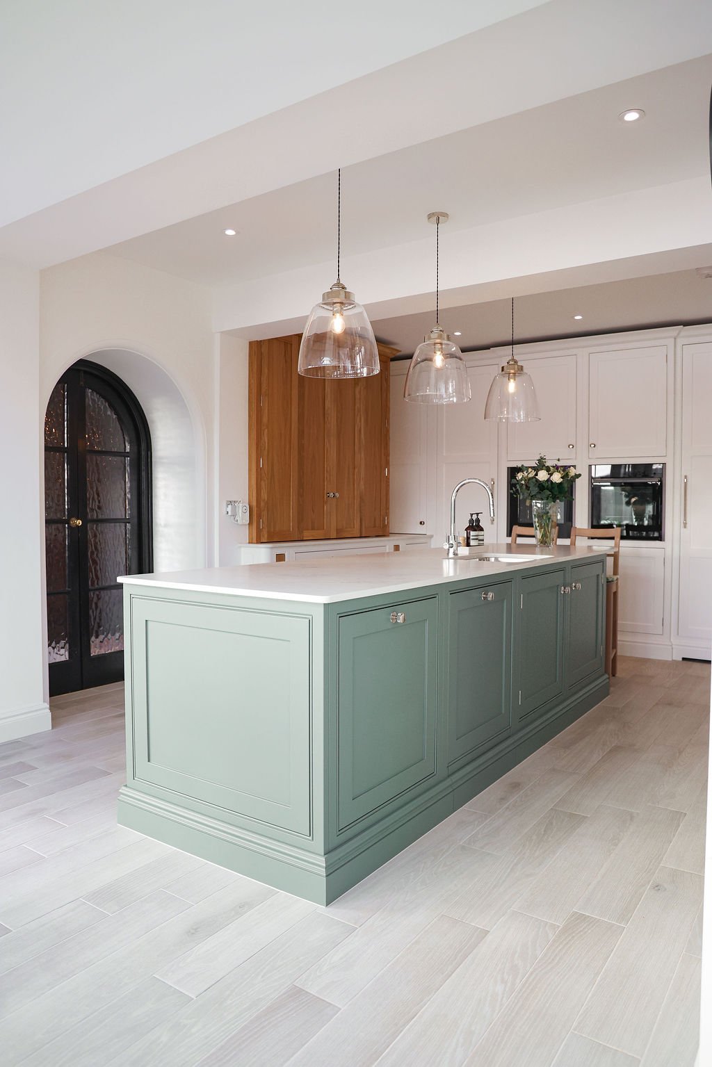 Modern kitchen with green island, white countertops, white cabinets, wooden accents, black arched door, pendant lights, and flowers on the counter.