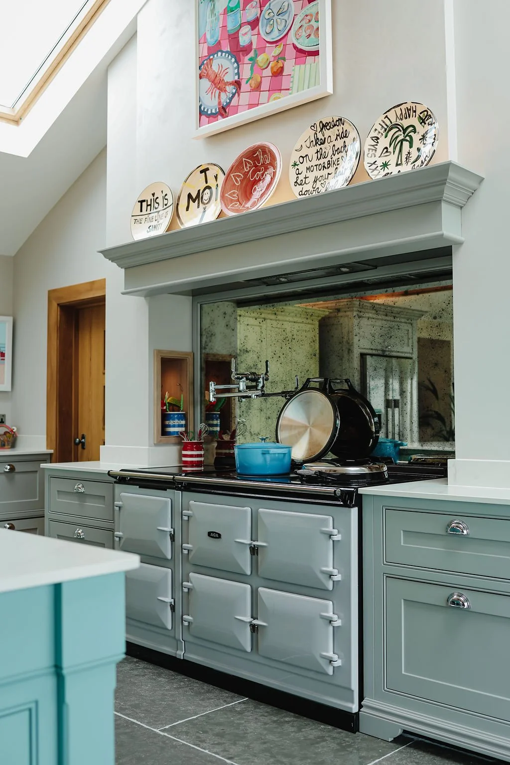 A vintage kitchen stove in light gray with multiple oven compartments, on a white countertop. Above it, decorative ceramic plates with writing and graphics are displayed on a white shelf. There is a large mirror behind the stove and colorful artwork on the wall. The kitchen features a skylight window on the sloped ceiling.