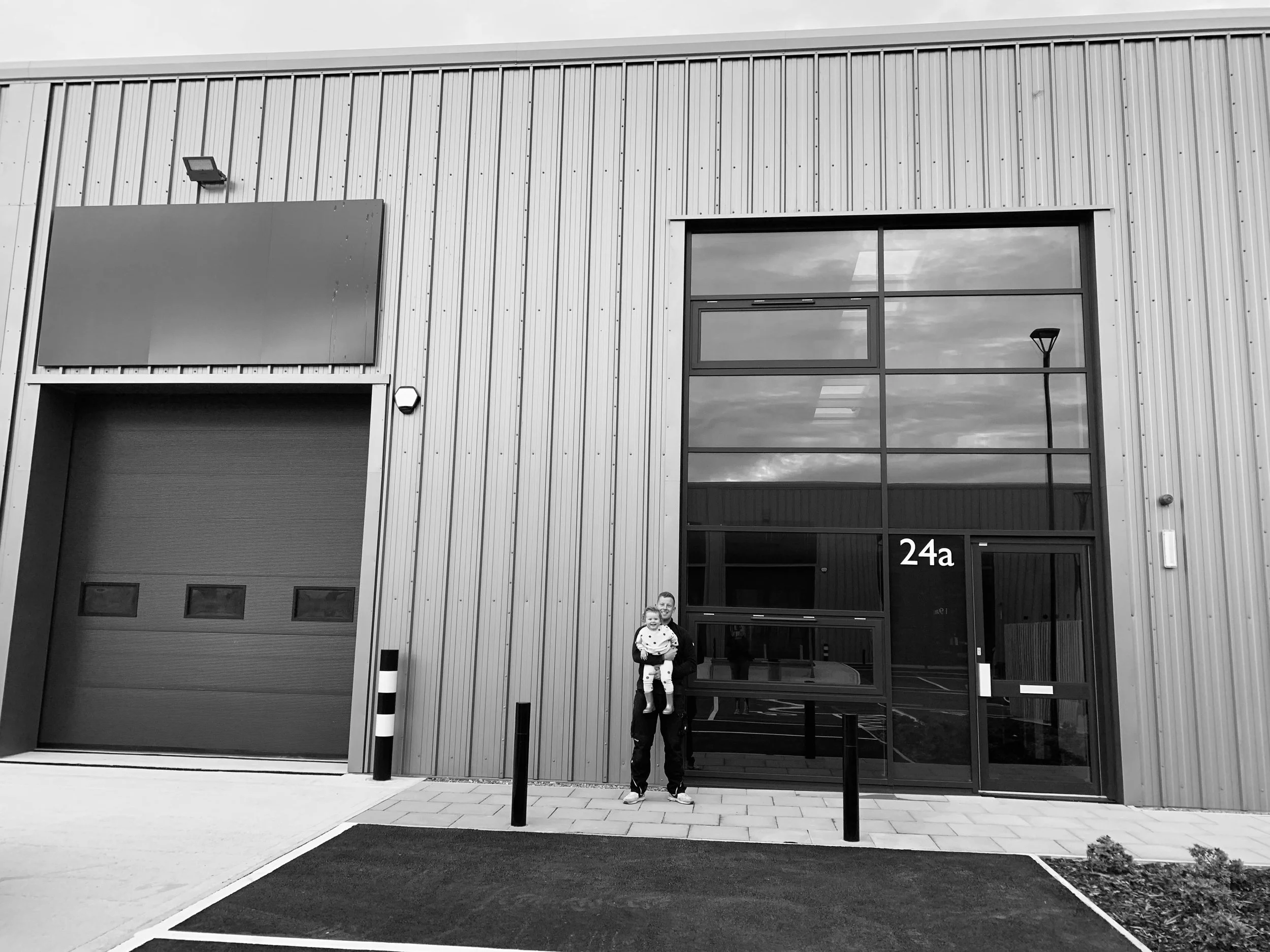 A person holding a child standing in front of a modern industrial building with large glass windows and a garage door, marked with the number 24a.