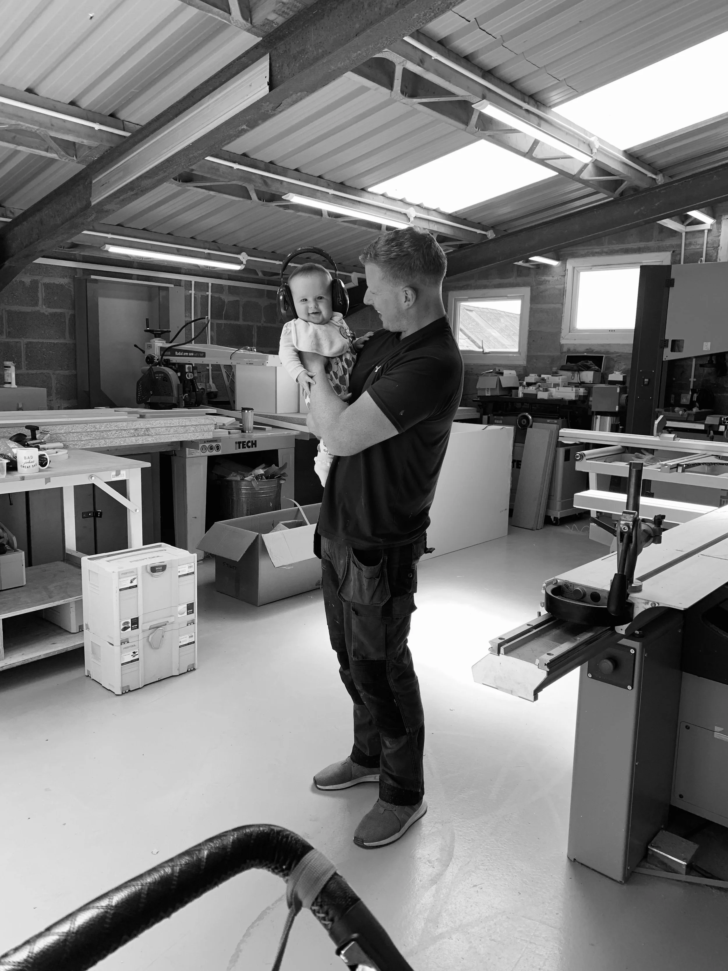A man holding a smiling baby girl with headphones in a woodworking workshop, surrounded by tools and equipment.