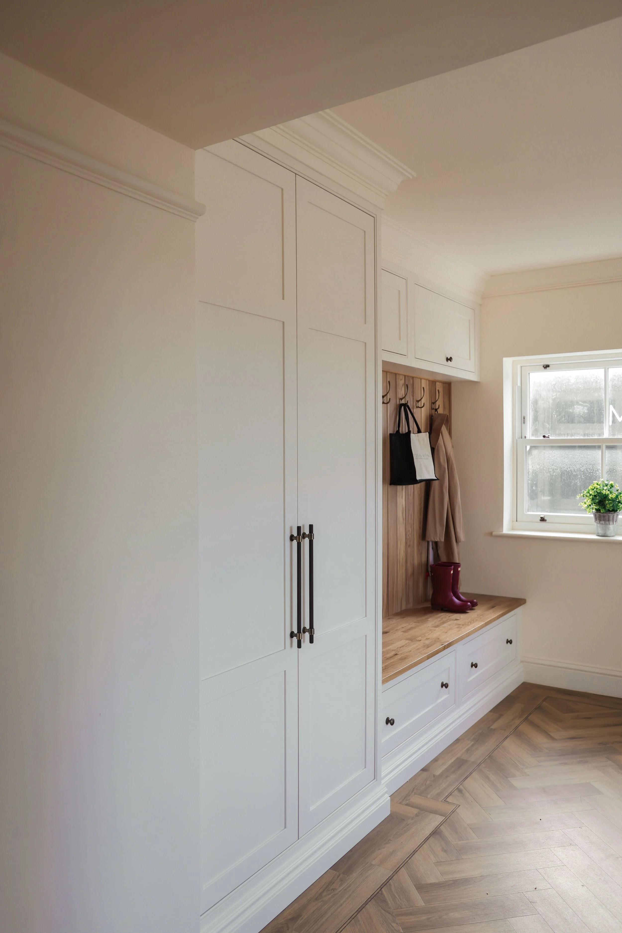 Corner of a home entryway with white built-in cabinets and a wooden bench beneath a window, decorated with a potted plant and coat hooks with bags and boots.