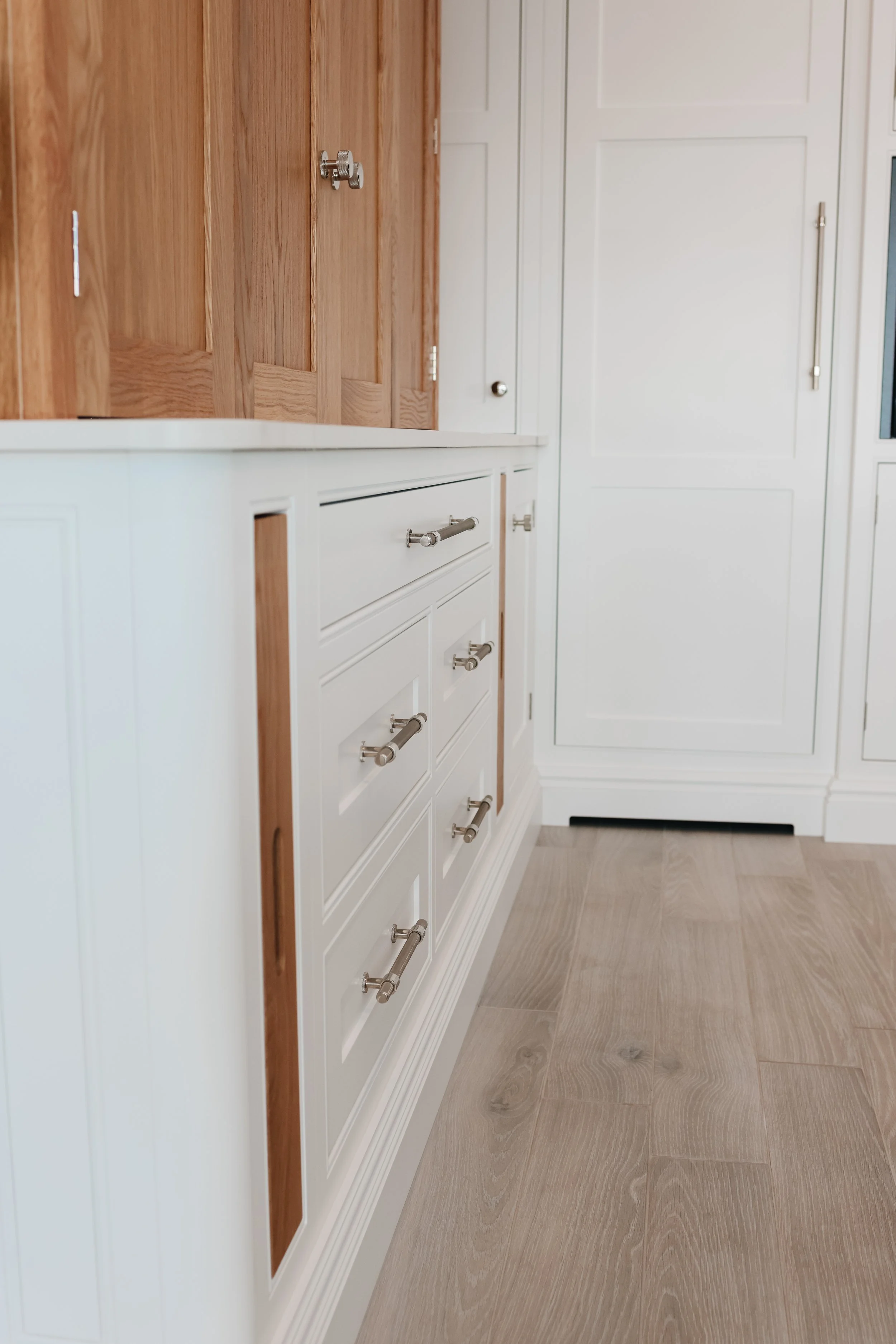 A close-up of kitchen cabinets, white on the lower section with silver handles, and wood-paneled on the upper section, with a wooden floor.