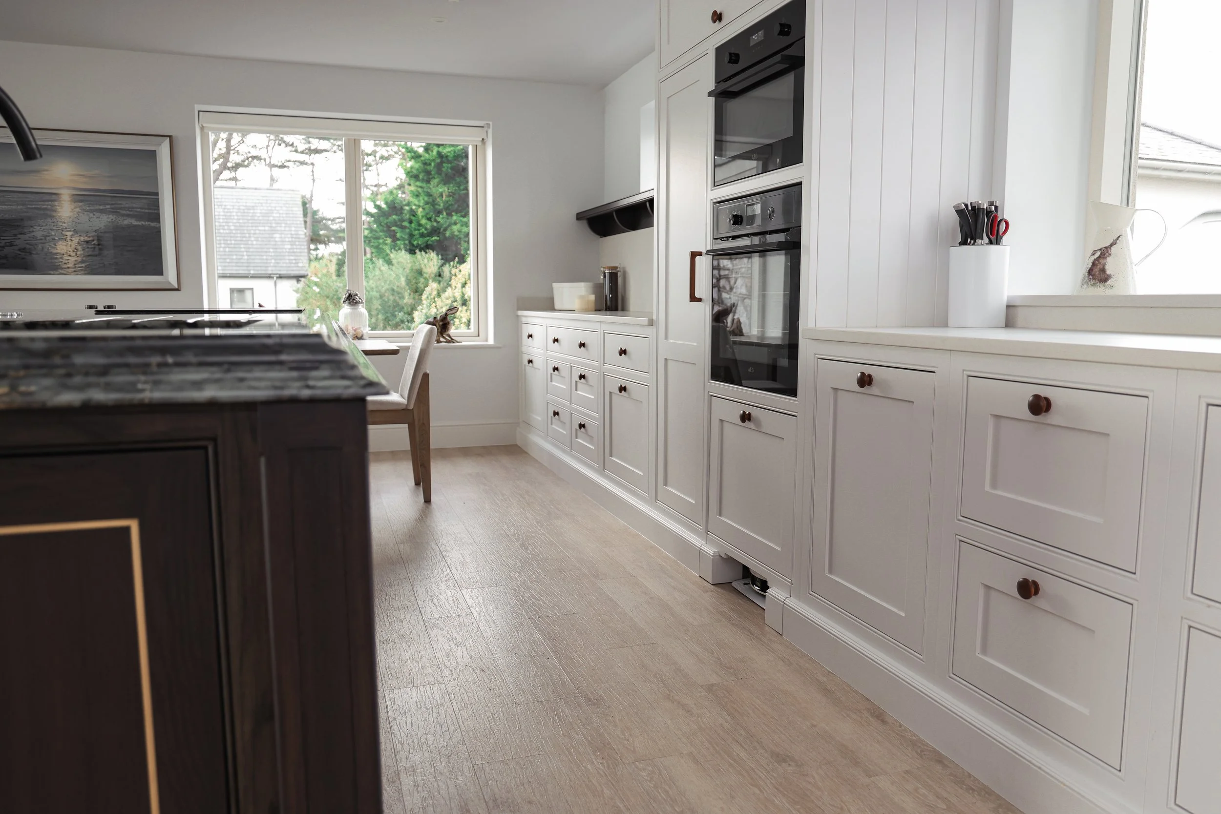 Bright modern kitchen with white cabinets, built-in oven, island with dark cabinetry, and a window overlooking green trees.