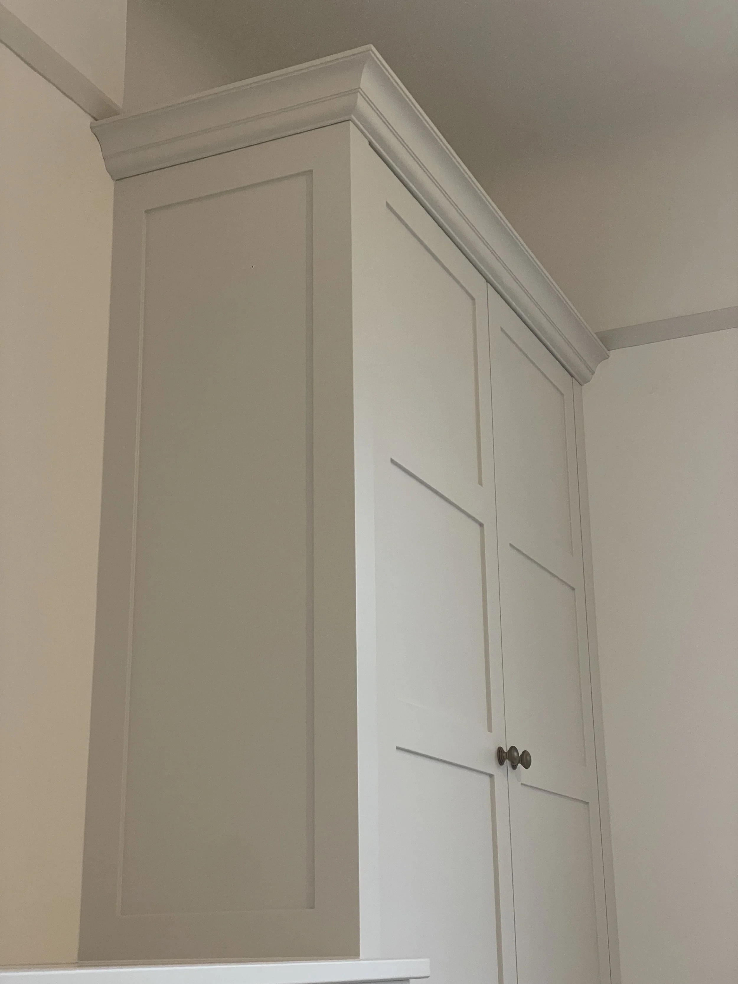 White corner cabinet with two doors and knobs, decorative crown molding at the top, against a light-colored wall and ceiling.