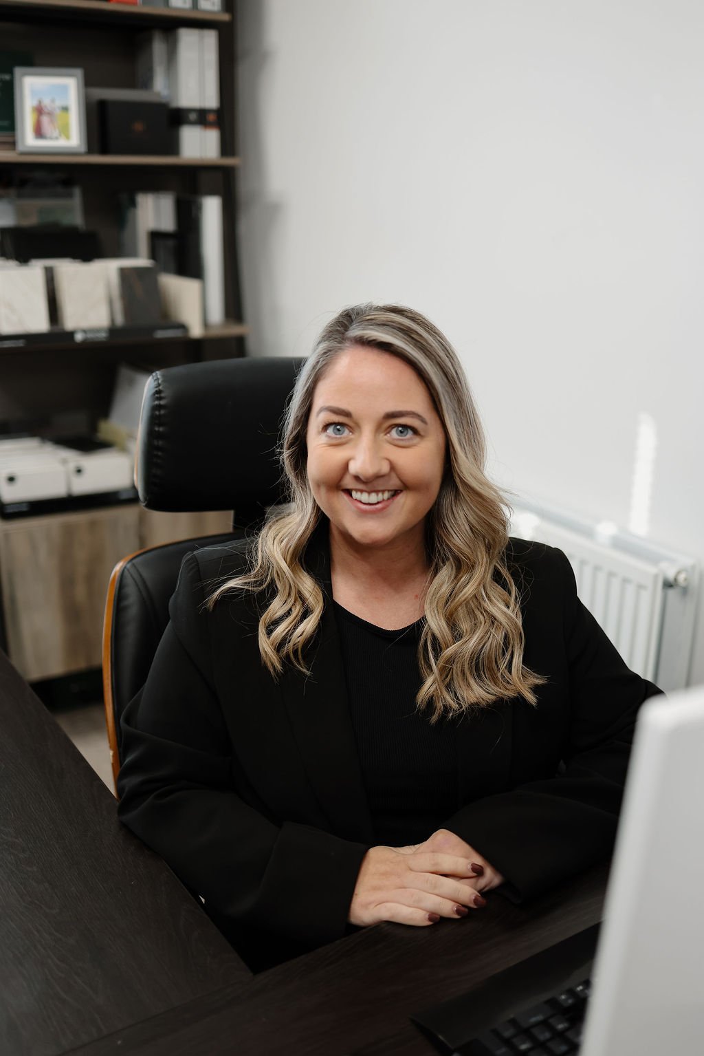 A smiling woman with long, wavy blonde hair sitting at a dark wooden desk in an office, wearing a black blazer.