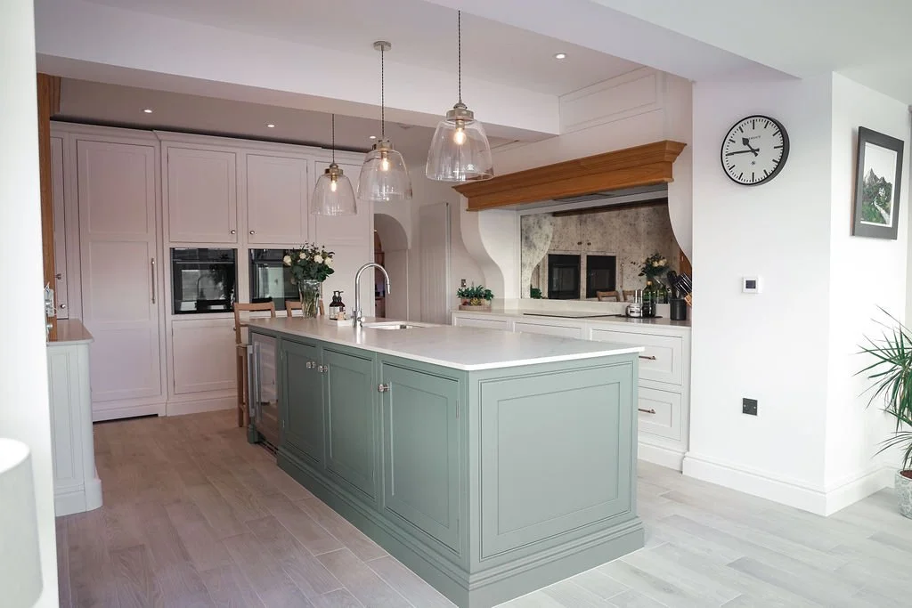 Modern kitchen with light-colored cabinets, a central green island, pendant lights, potted plants, and a wall clock.