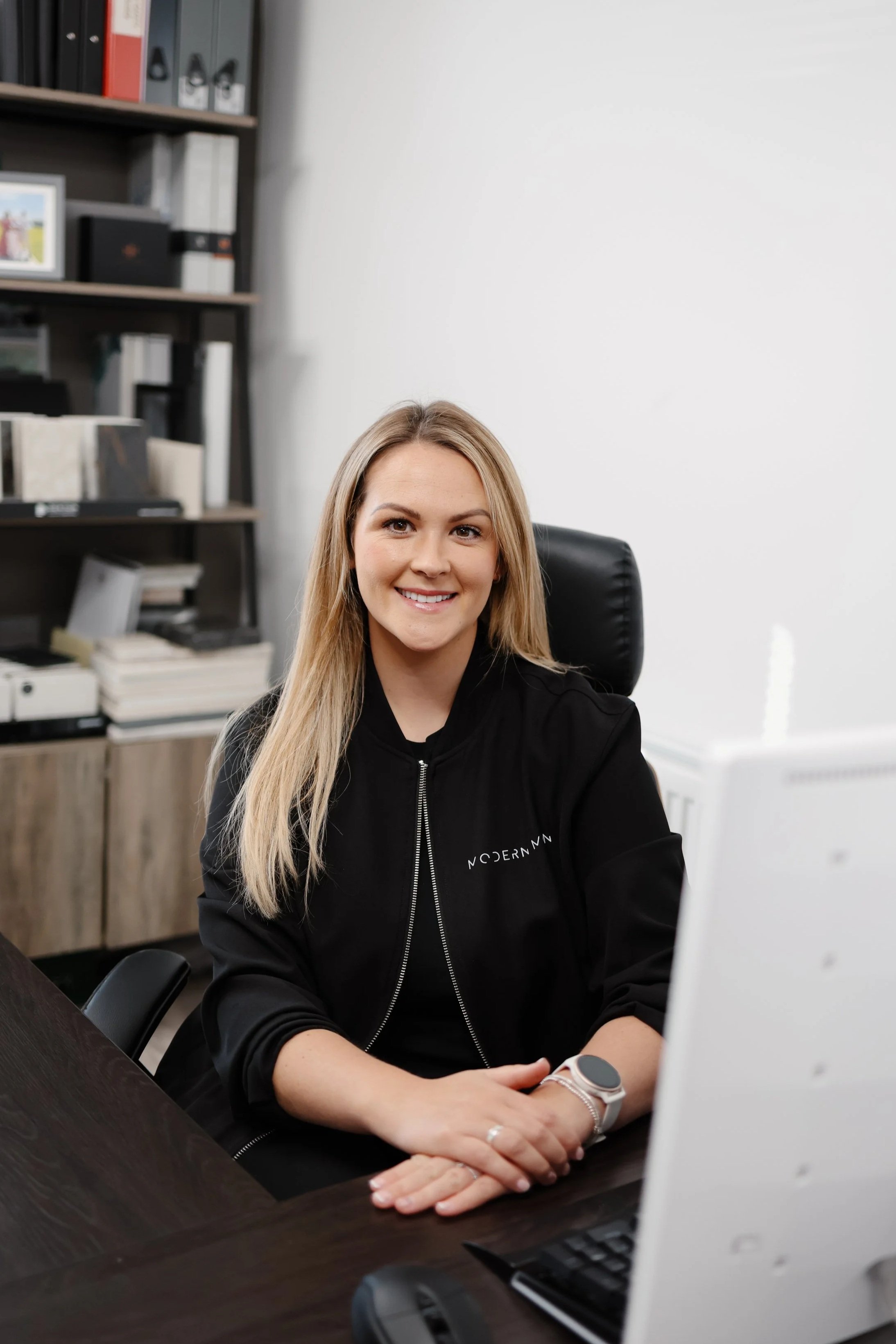A young woman with long blonde hair sitting at a desk in an office, smiling at the camera, wearing a black jacket with the word 'MODERNN' on it, with shelves full of books and binders in the background.