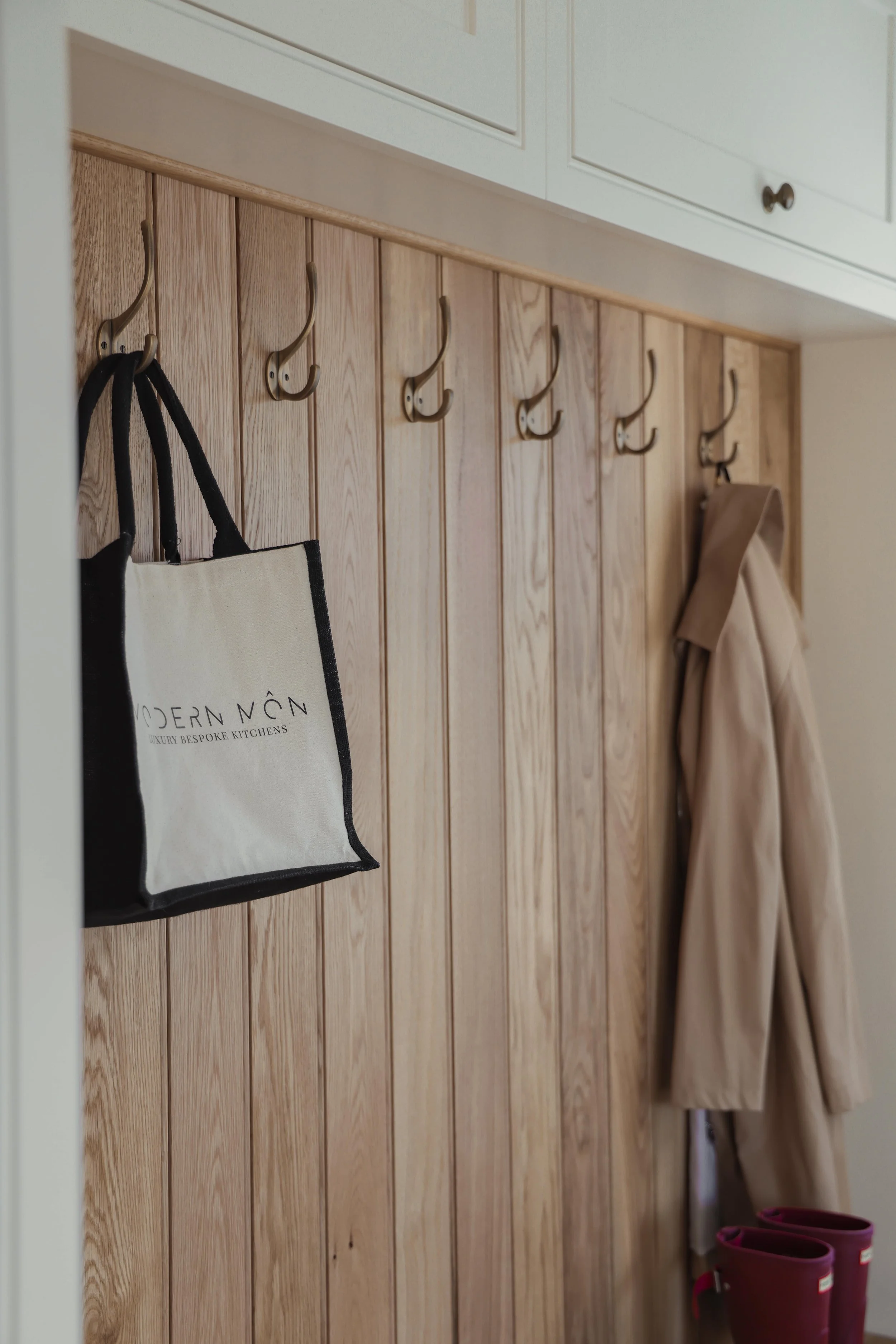 Interior of a mudroom with wooden paneling, gold hooks on the wall, a beige coat hanging, a canvas tote bag on one of the hooks, and pink rain boots on the floor.