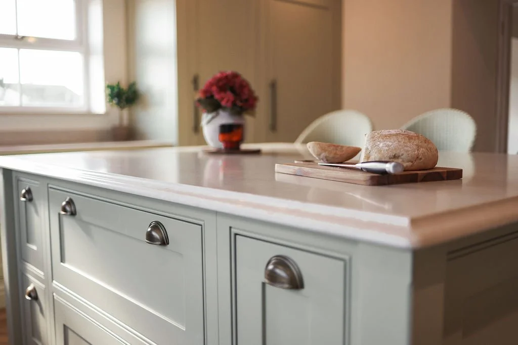 A kitchen island with a light-colored countertop, silver cabinet handles, and a flower vase with pink flowers in the background.
