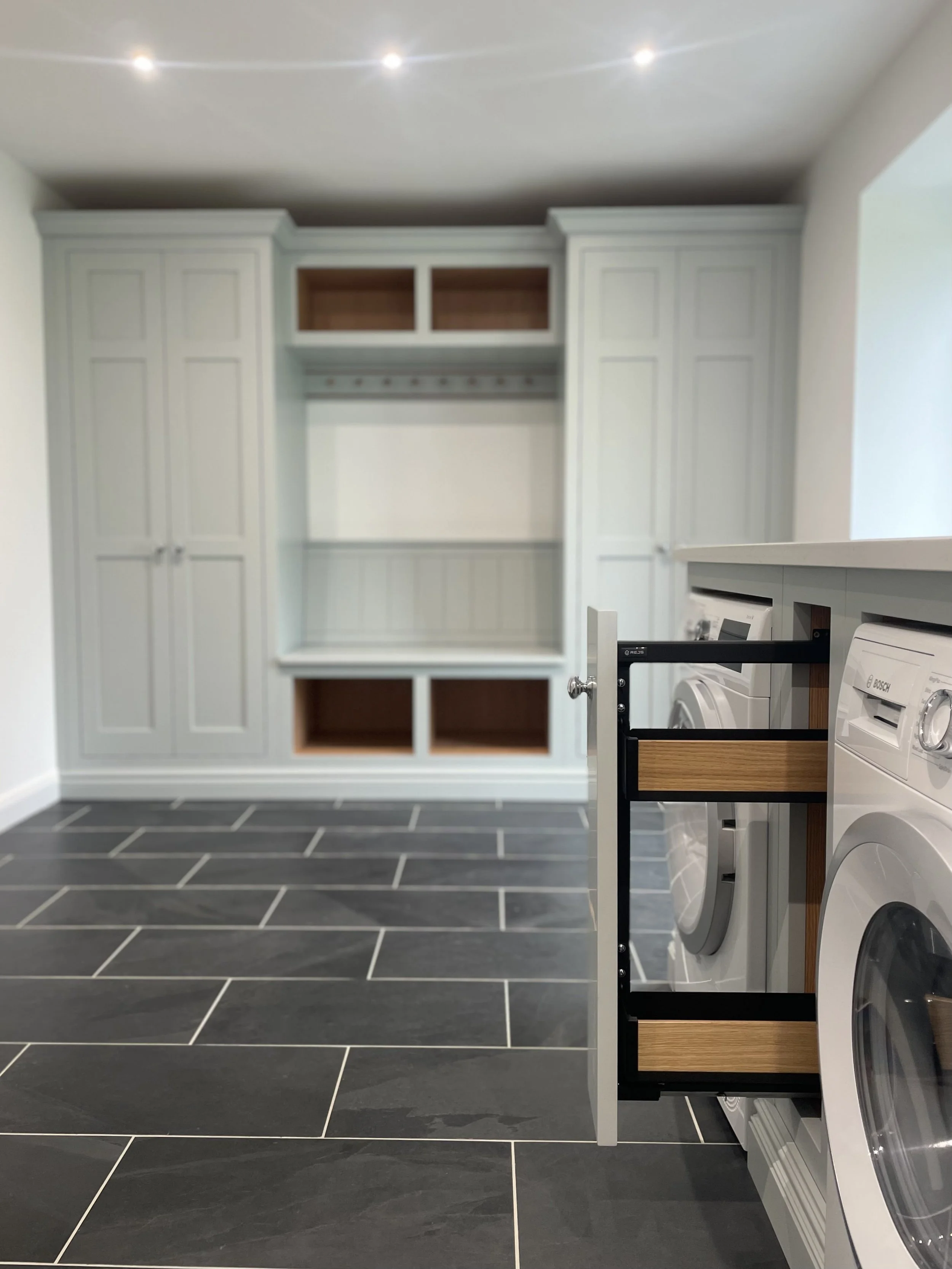 Modern laundry room with white cabinetry, dark tiled floor, and laundry appliances, including a washing machine and dryer, partially pulled out from a cabinet.