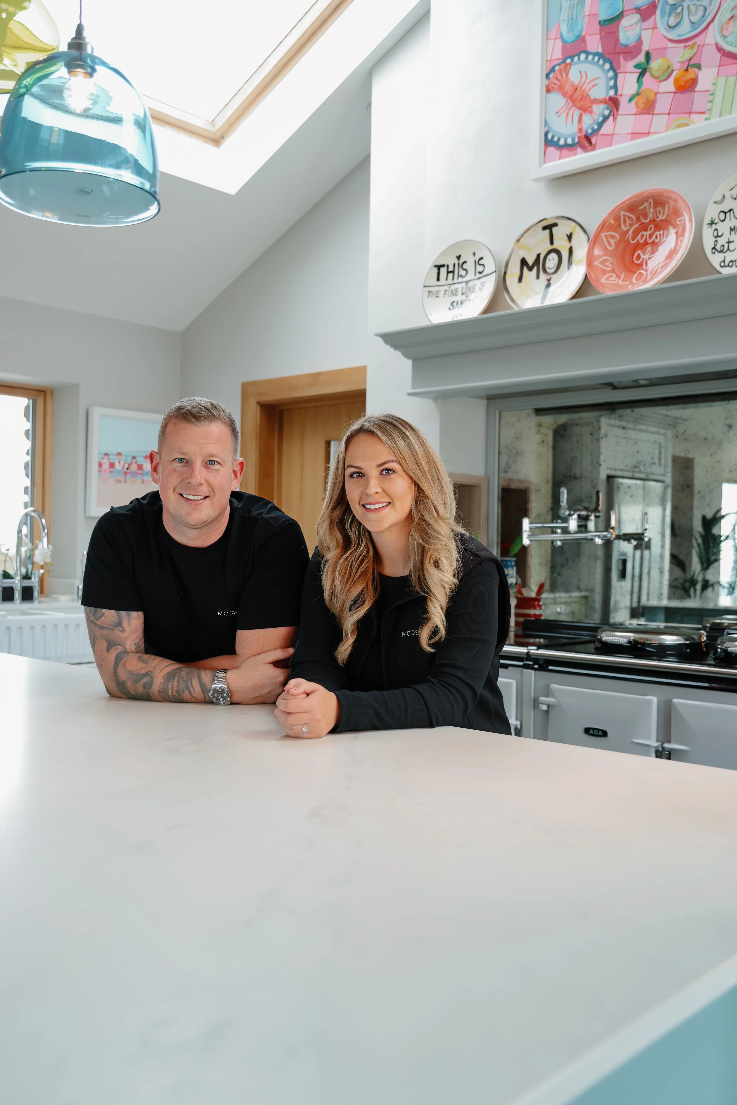A man and a woman sitting at a kitchen counter, smiling at the camera. The man has tattoos on his arm and short blond hair, while the woman has long blond hair and is wearing a black jacket. The kitchen is bright and modern, with decorative plates on the shelf above the stovetop and a colorful painting on the wall.