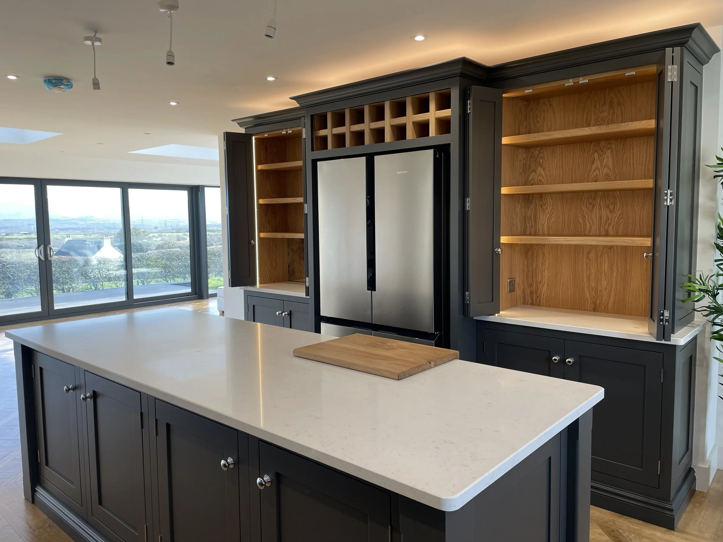 Modern kitchen with dark gray cabinets, white countertop island, stainless steel refrigerator, open shelving, large windows, and a scenic view.